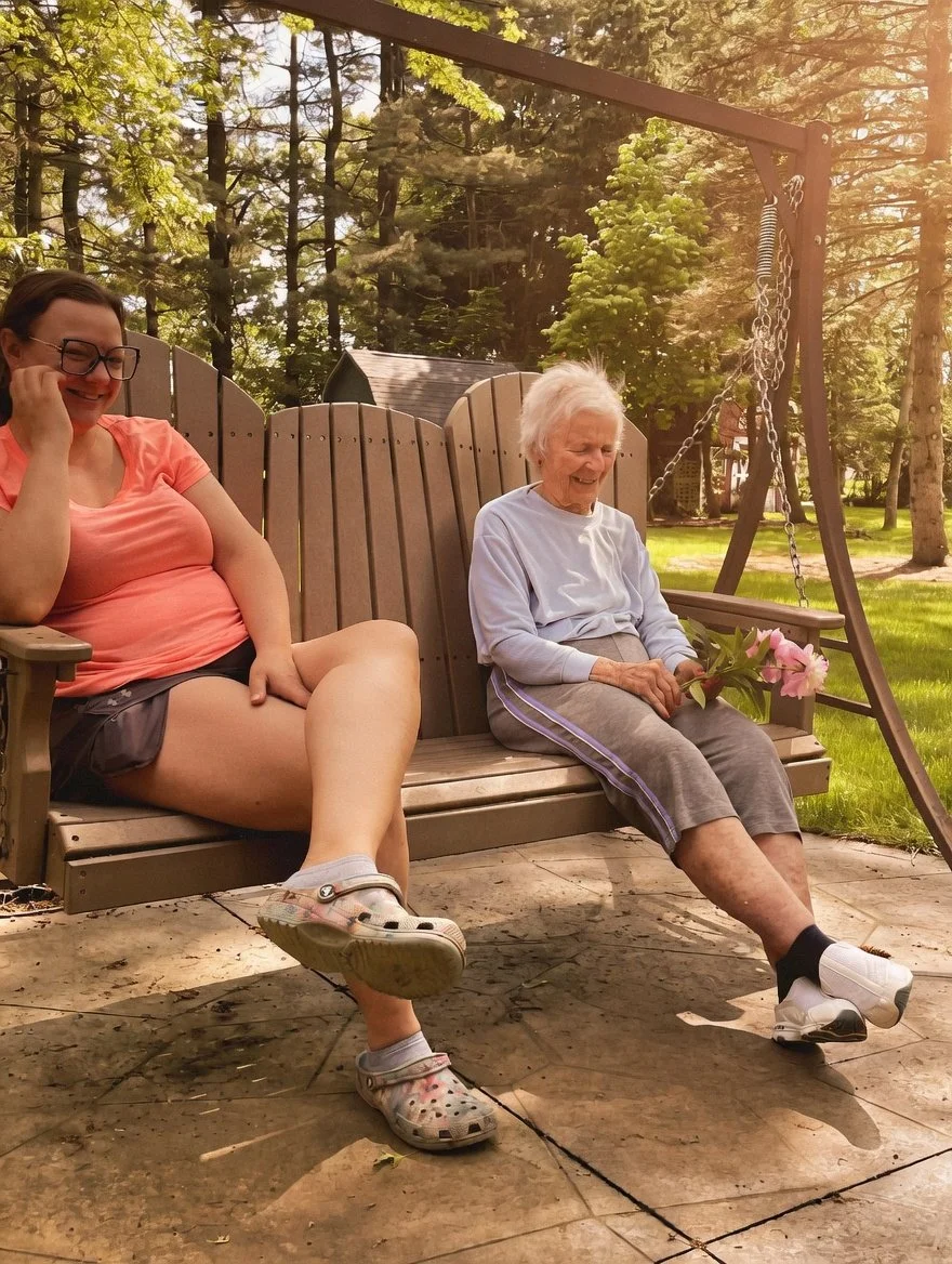 An elderly woman sitting on a wooden swing bench holding pink flowers, smiling, with a younger woman sitting beside her on the same swing. They are outdoors in a park with trees and sunlight.