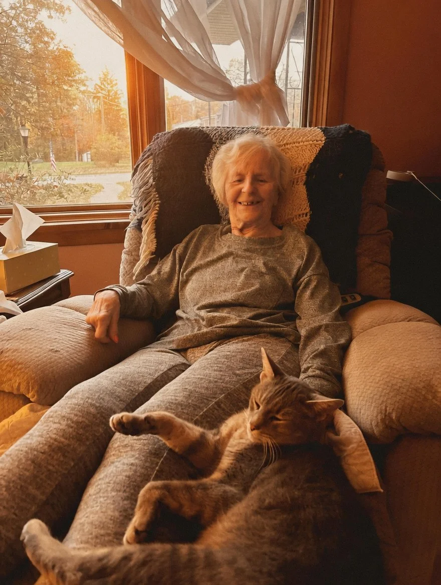 An elderly woman sitting on a beige armchair with a patterned blanket behind her, smiling at the camera with a gray tabby cat lying on her lap, in a room with sunlight coming through a window with sheer curtains.
