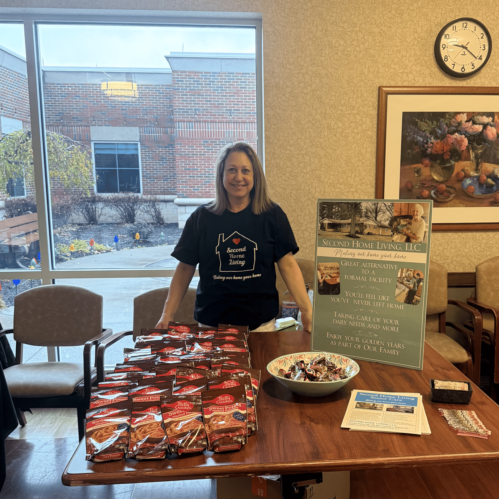 A woman standing behind a table with packets of Betty Crocker sweet potatoes and a bowl of individually wrapped candies. There are chairs, a window, and a promotional poster for second home living and senior care in the background.