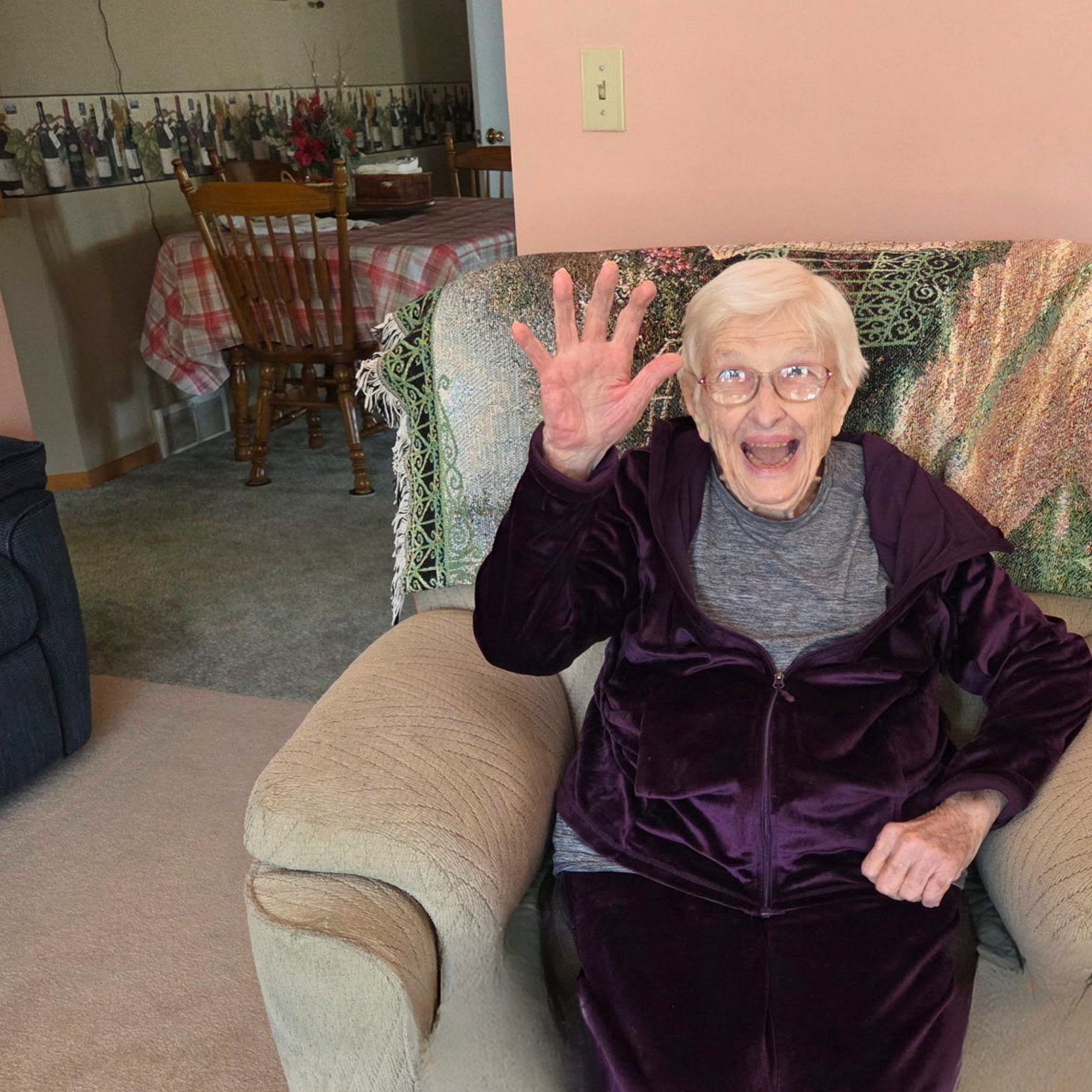 An elderly woman with short white hair and glasses sitting on a beige armchair. She is smiling and waving with her right hand, wearing a gray shirt and a purple velvet jacket. Behind her is a patterned blanket over the back of the chair and pink painted wall.