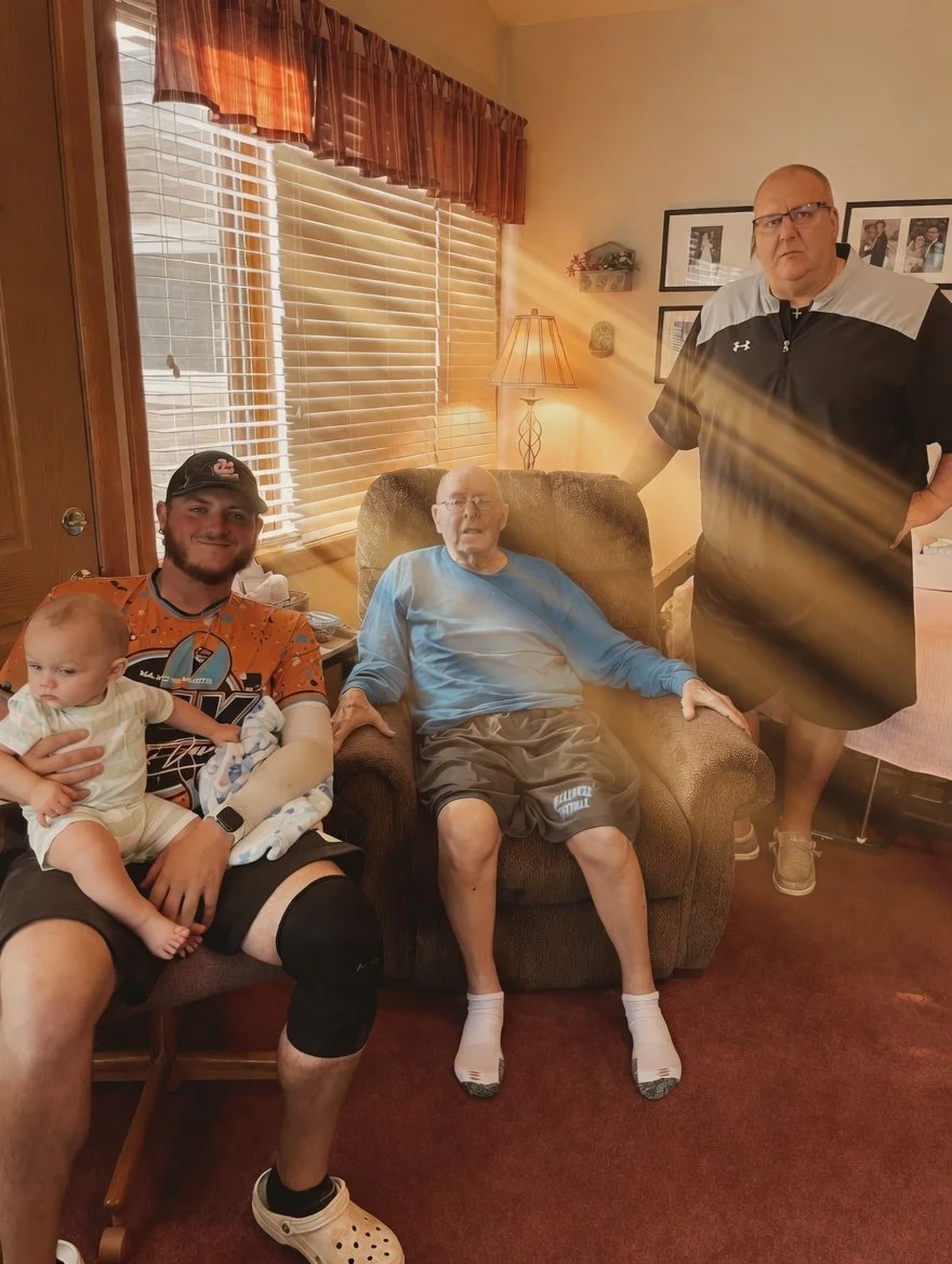 A family gathering in a living room with four people, including a baby, an elderly man seated in a recliner, a young man holding the baby, and a middle-aged man standing. The room has wooden blinds, a lamp, framed photos on the wall, and a window wit