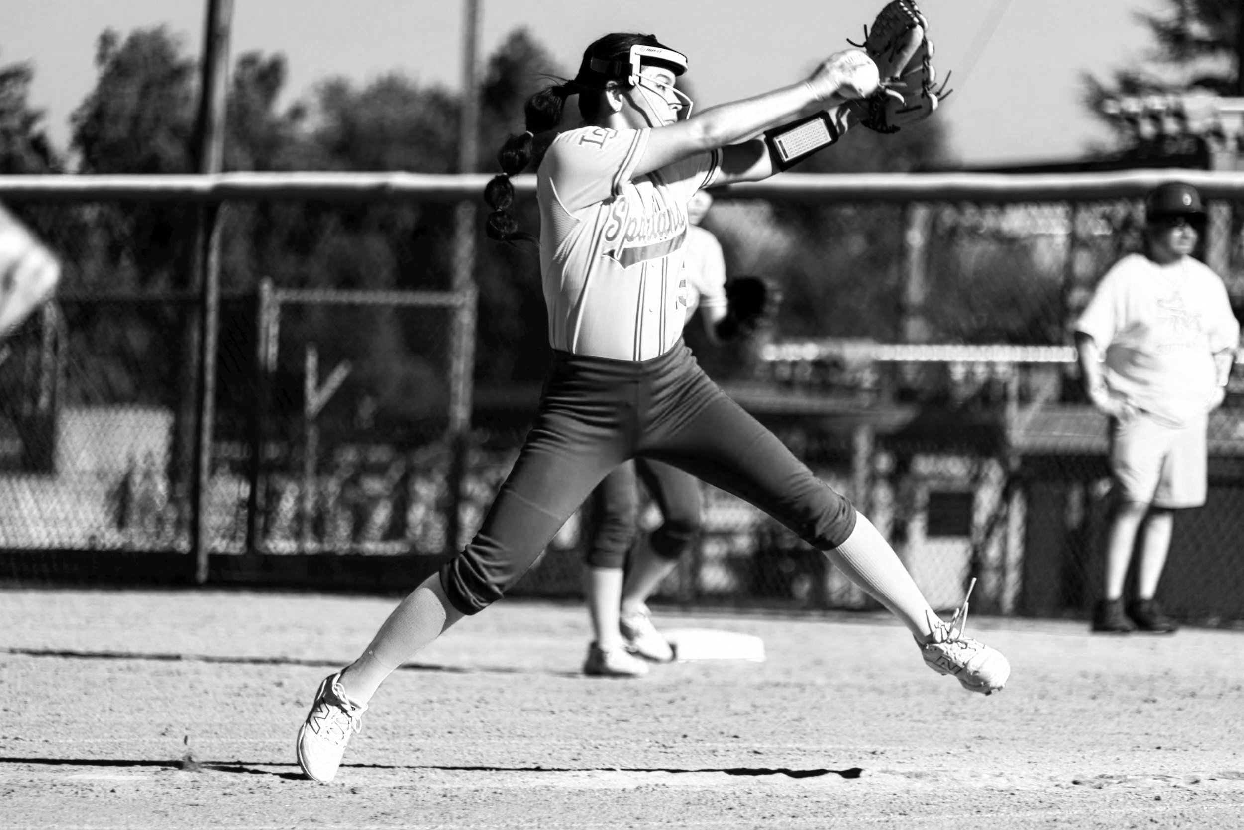 A female softball player in uniform stretching before a game, wearing protective goggles and holding a glove, with a teammate in the background on the field.