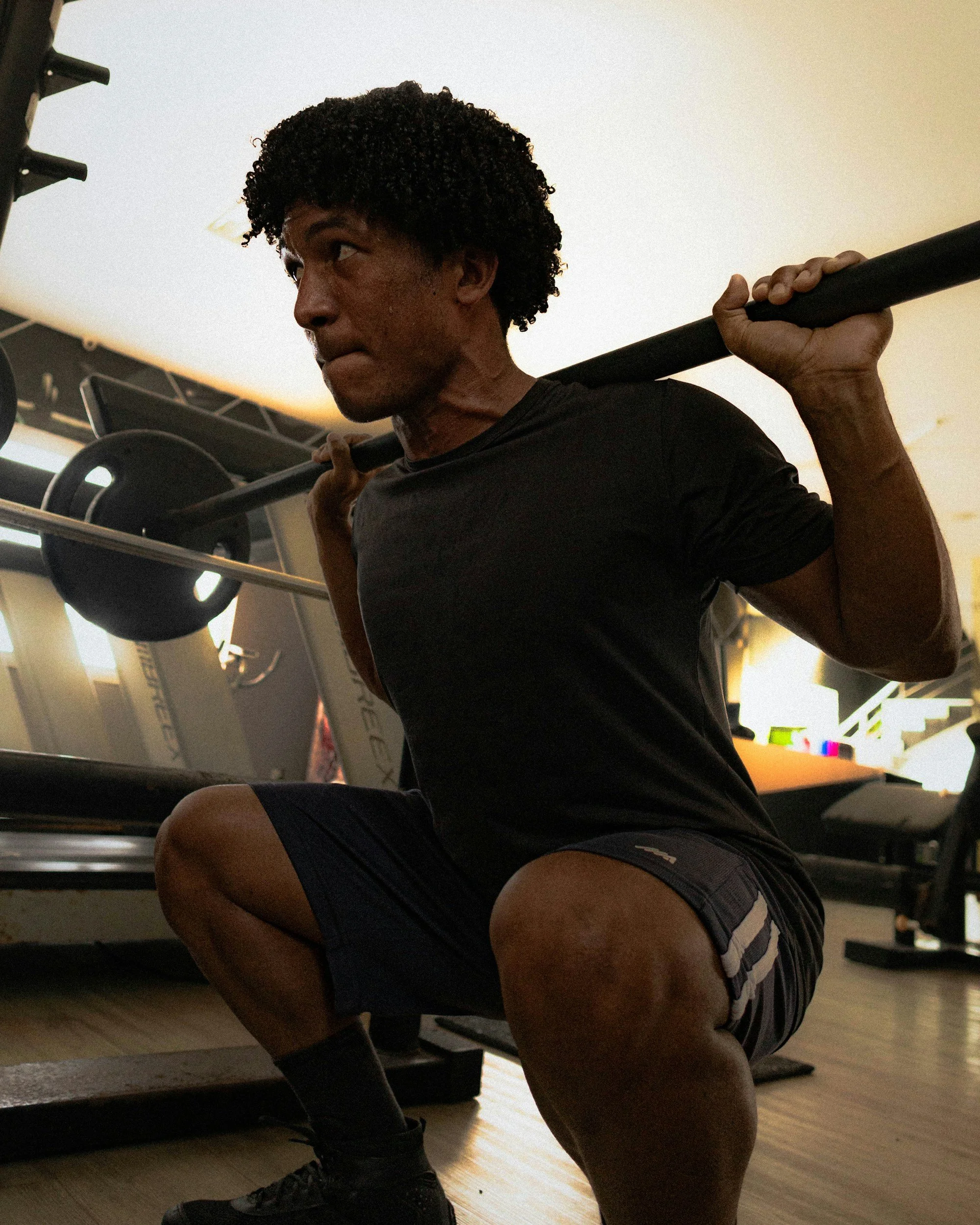 A man with curly black hair in a gym squat position, holding a barbell across his shoulders, wearing a black shirt, shorts, and black athletic shoes.