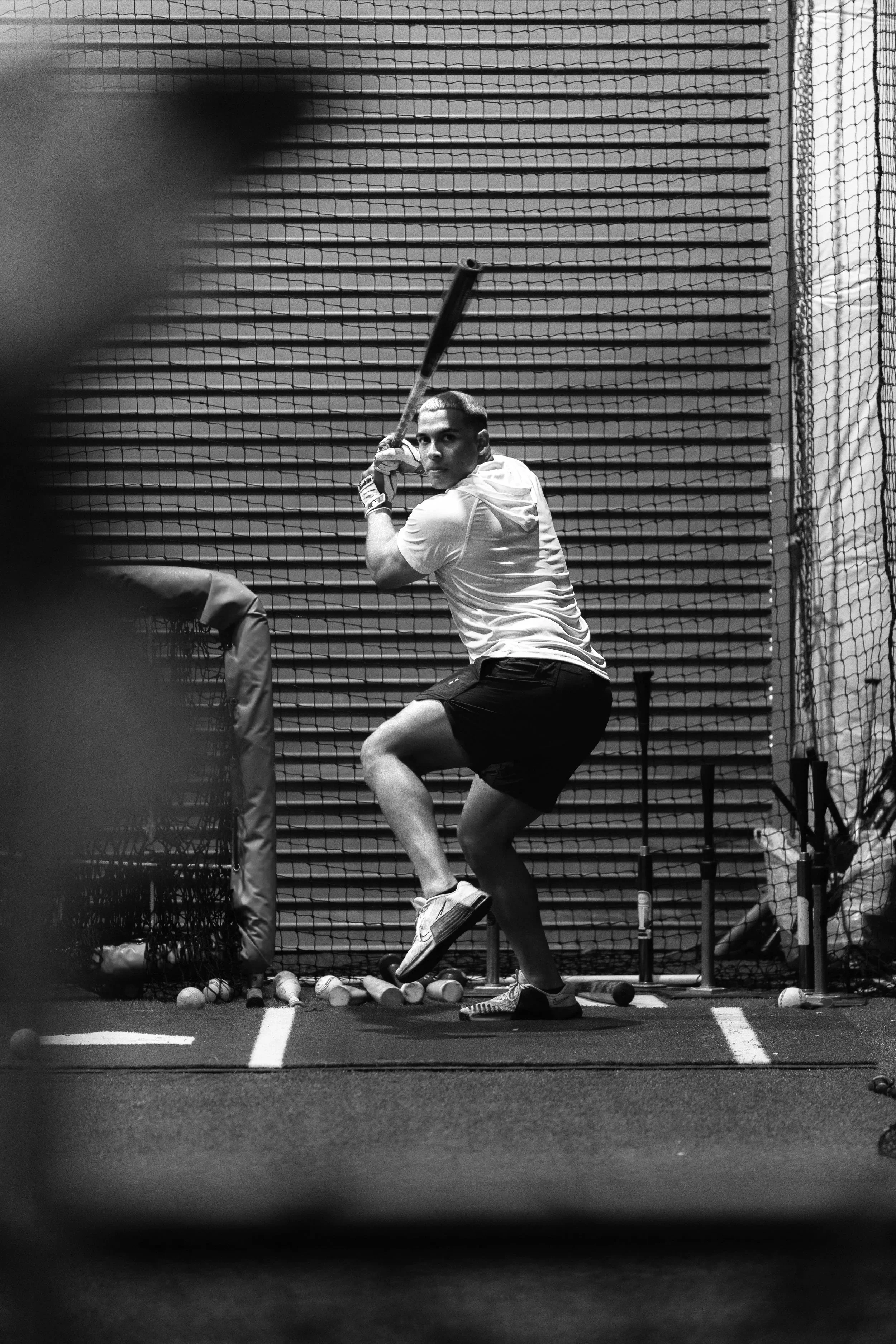 A young man in a white shirt and shorts practicing baseball batting inside a batting cage. He is holding a baseball bat, preparing to swing. Baseballs are scattered on the floor around him. The background features a wire mesh fence and horizontal slats.