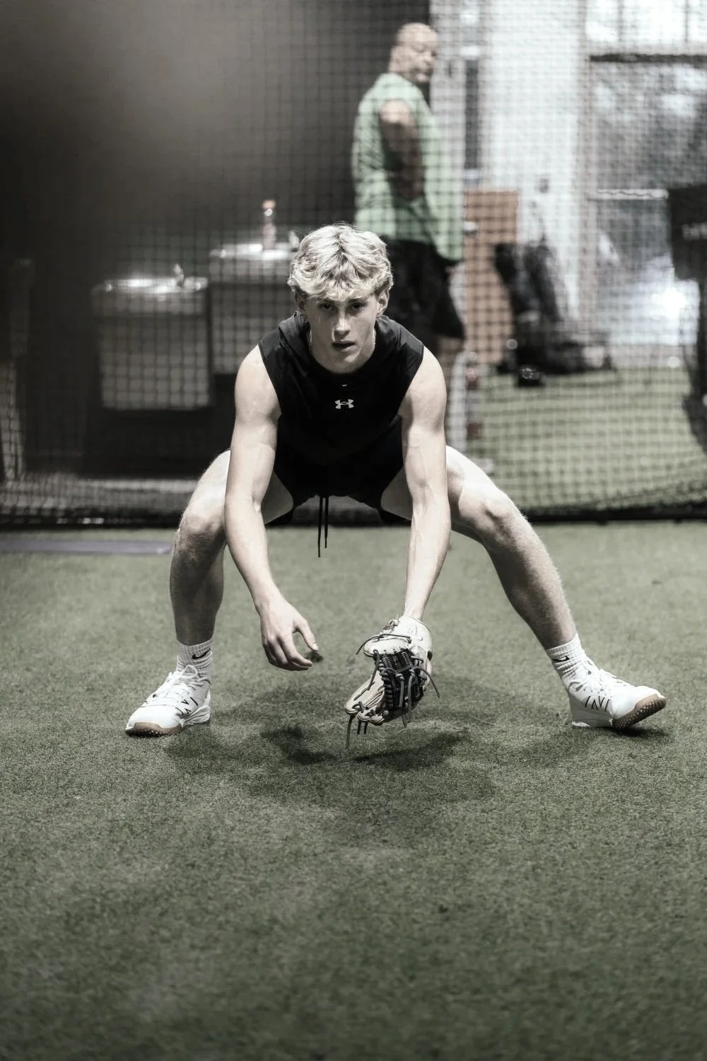 A young male athlete in a crouched ready position on a turf field, wearing athletic clothing and shoes, holding a baseball glove, with a netted enclosure and other people in the background