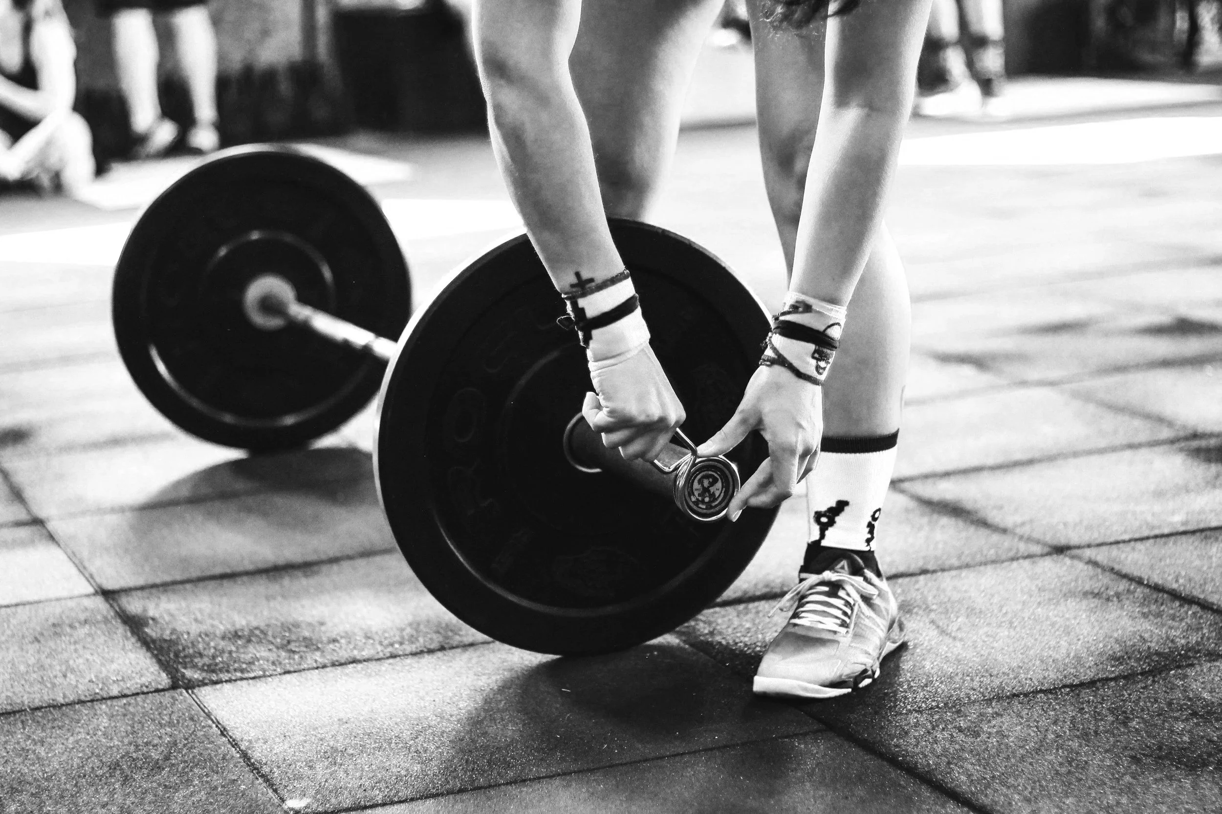 A person in athletic shoes and socks is lifting a barbell with weights in a gym, with other people in the background.