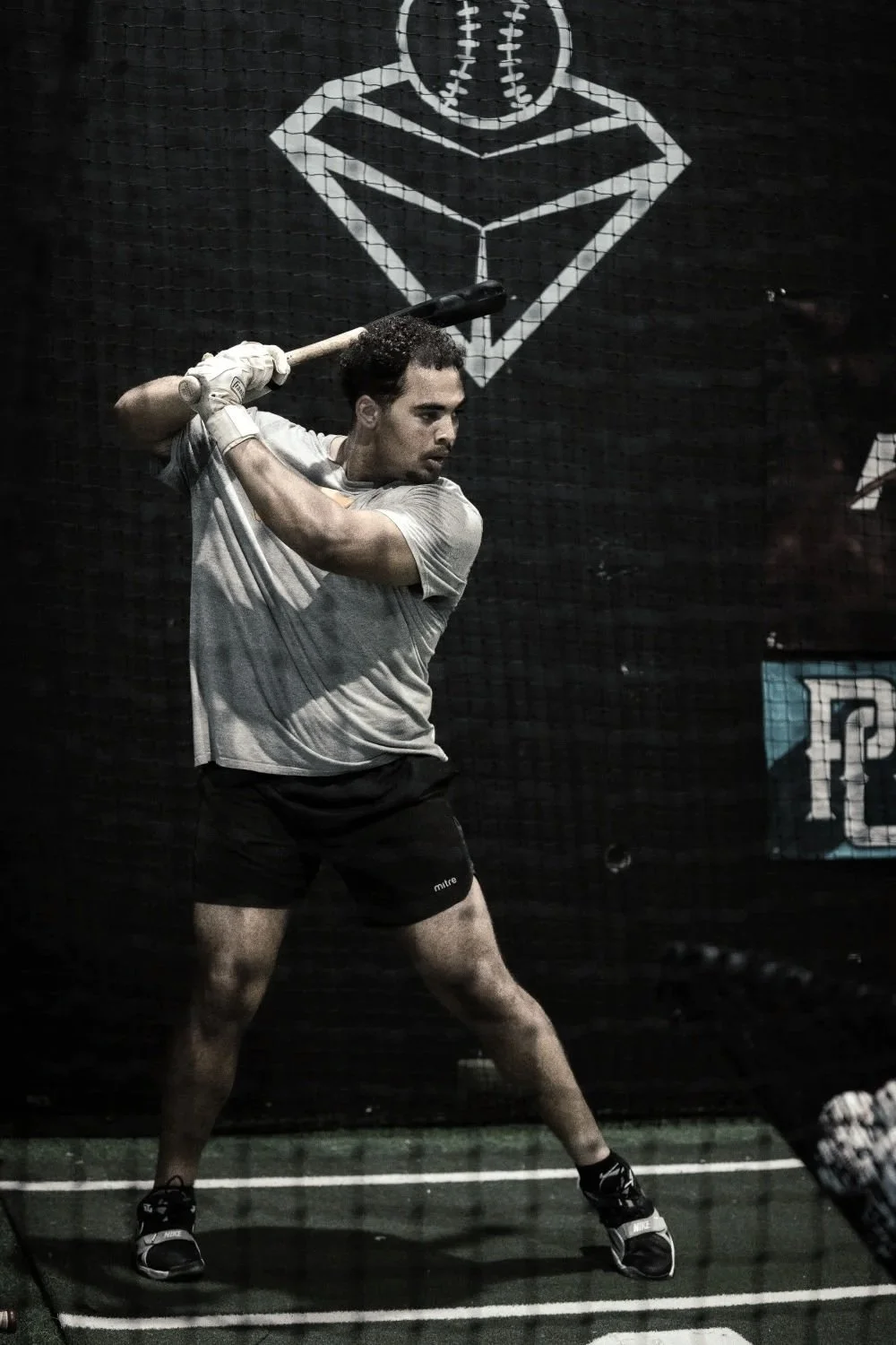 Man swinging a baseball bat indoors, with a logo of a baseball in a diamond behind him.