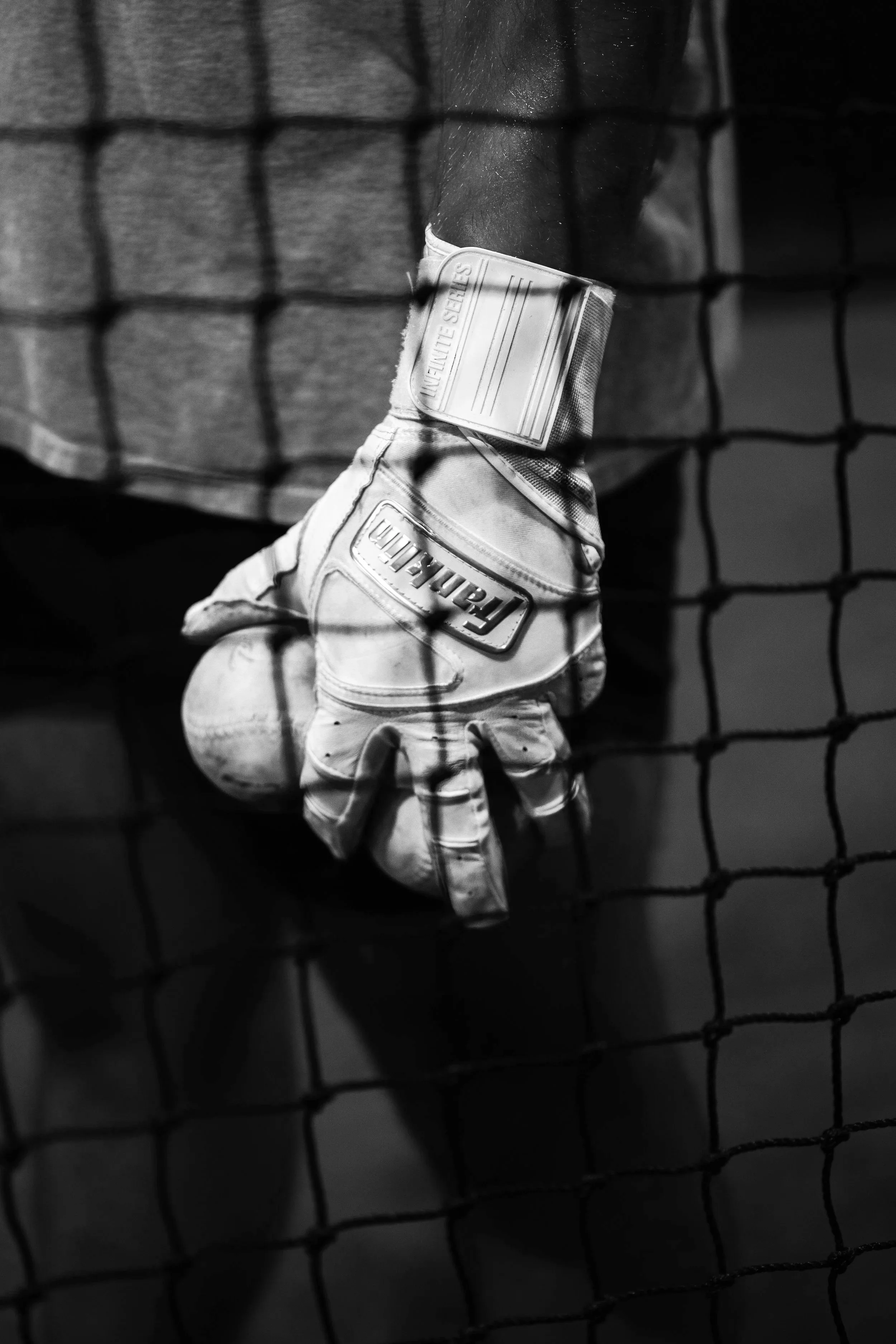 A black and white photograph taken through a wire fence showing a person wearing a glove, possibly a sports glove, with the word 'LEATHER' visible. The person is also wearing a textured shirt or jacket.
