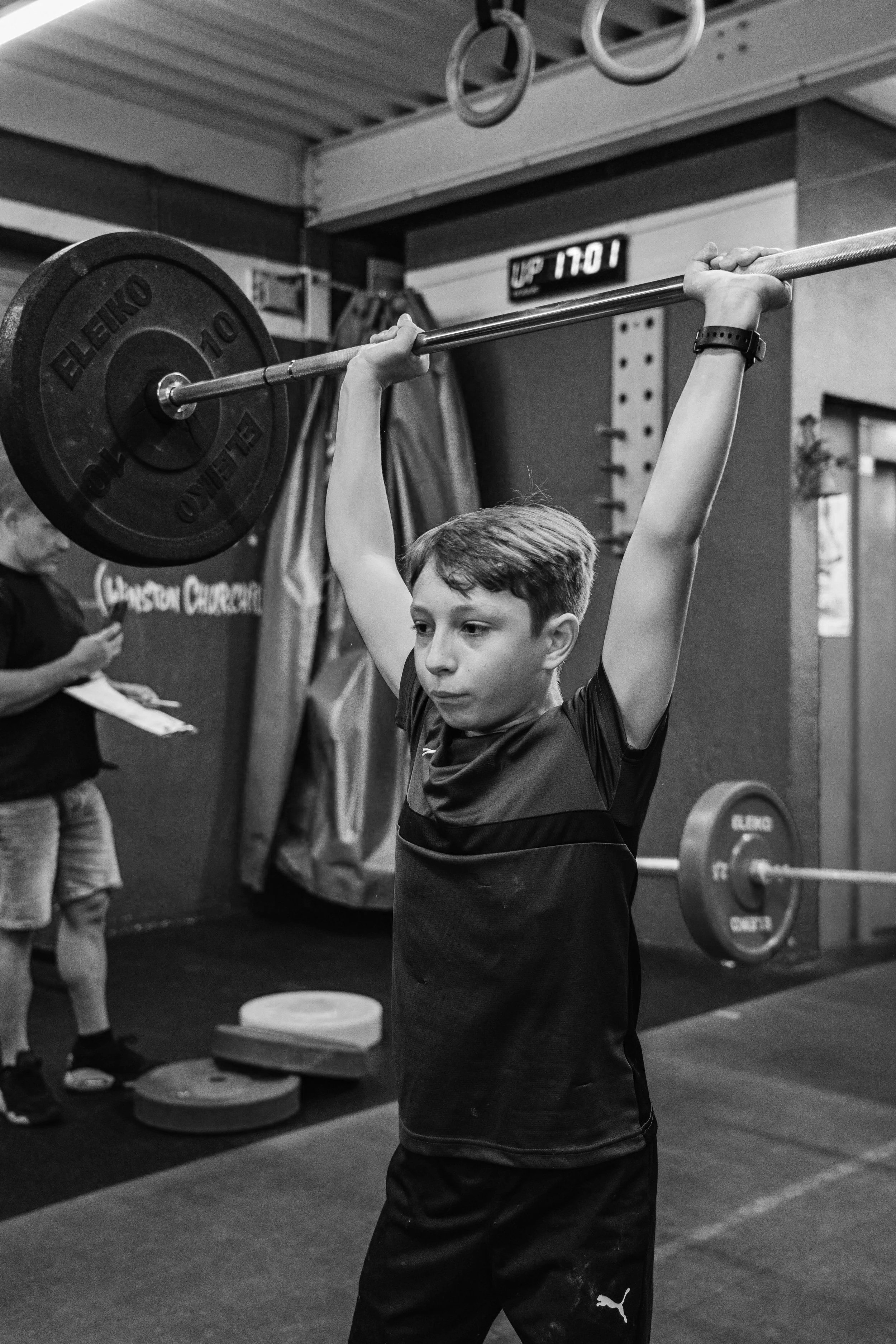 A boy lifting a barbell overhead in a gym, with weight plates on both ends, while a woman looks on in the background.