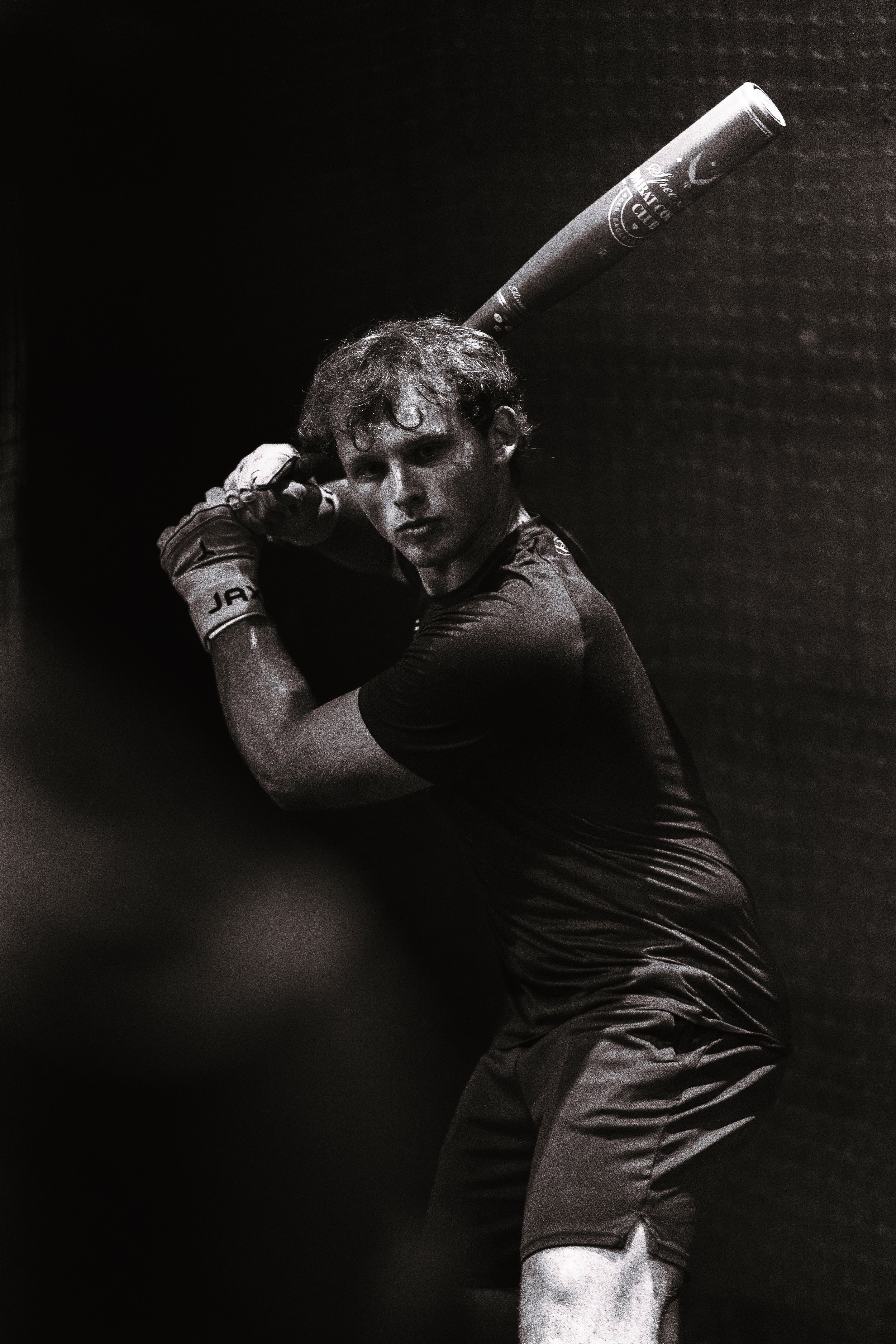 A young man dressed in athletic clothing is holding a baseball bat over his shoulder in a batting stance, looking towards the camera against a dark background.