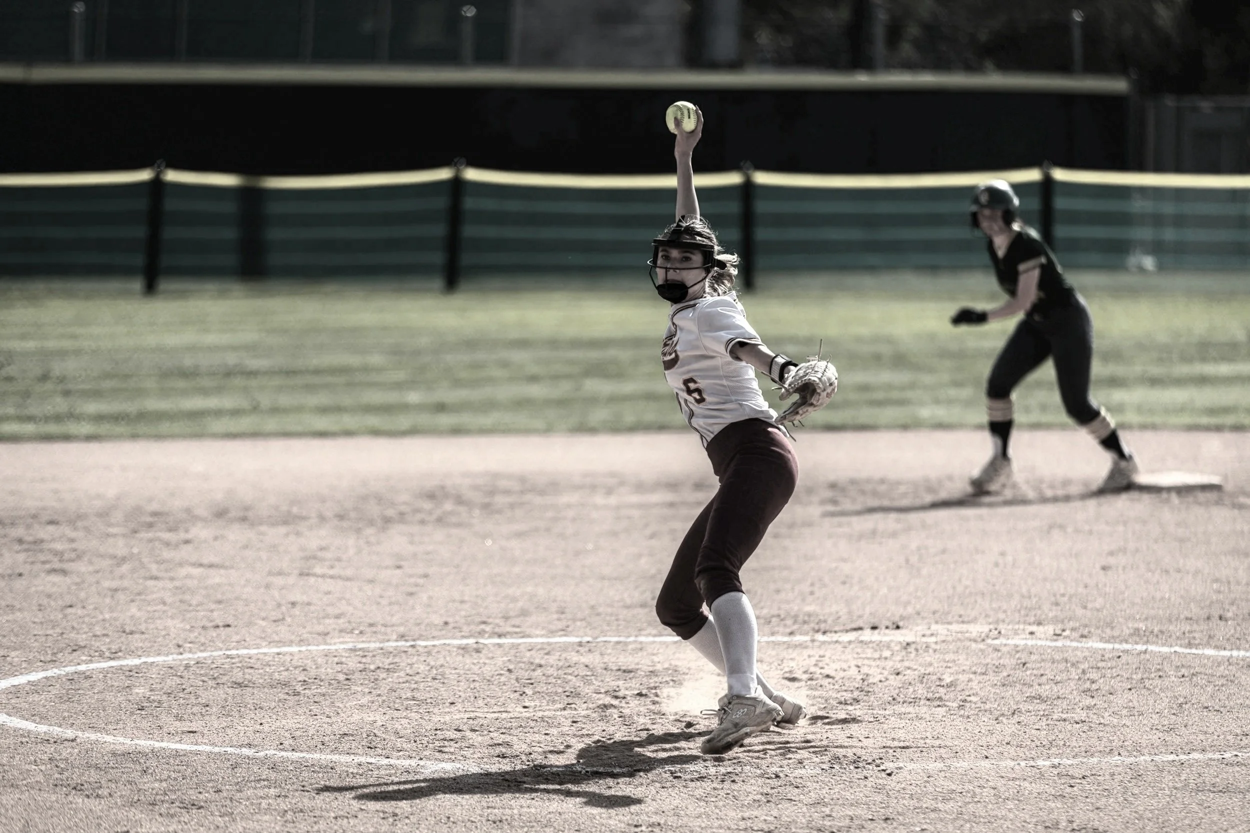 A young girl playing softball on a field, wearing a helmet and holding a baseball glove, with her arm raised preparing to catch or throw the ball, and another player in the background.