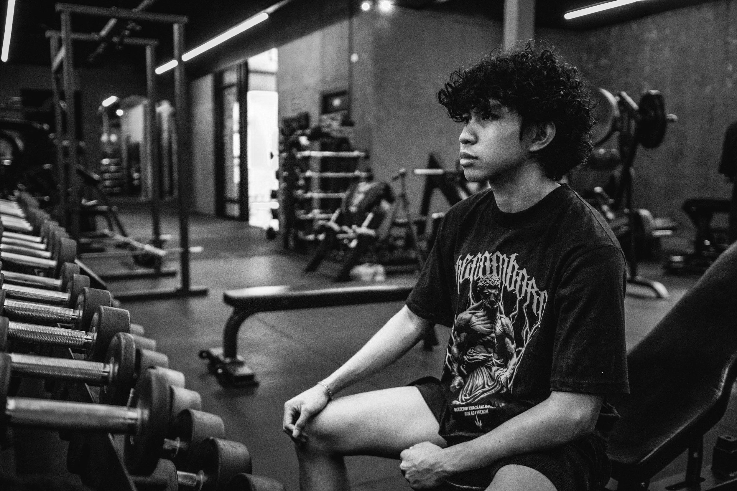 A young man with curly hair sitting on a bench in a gym, with weightlifting equipment in the background