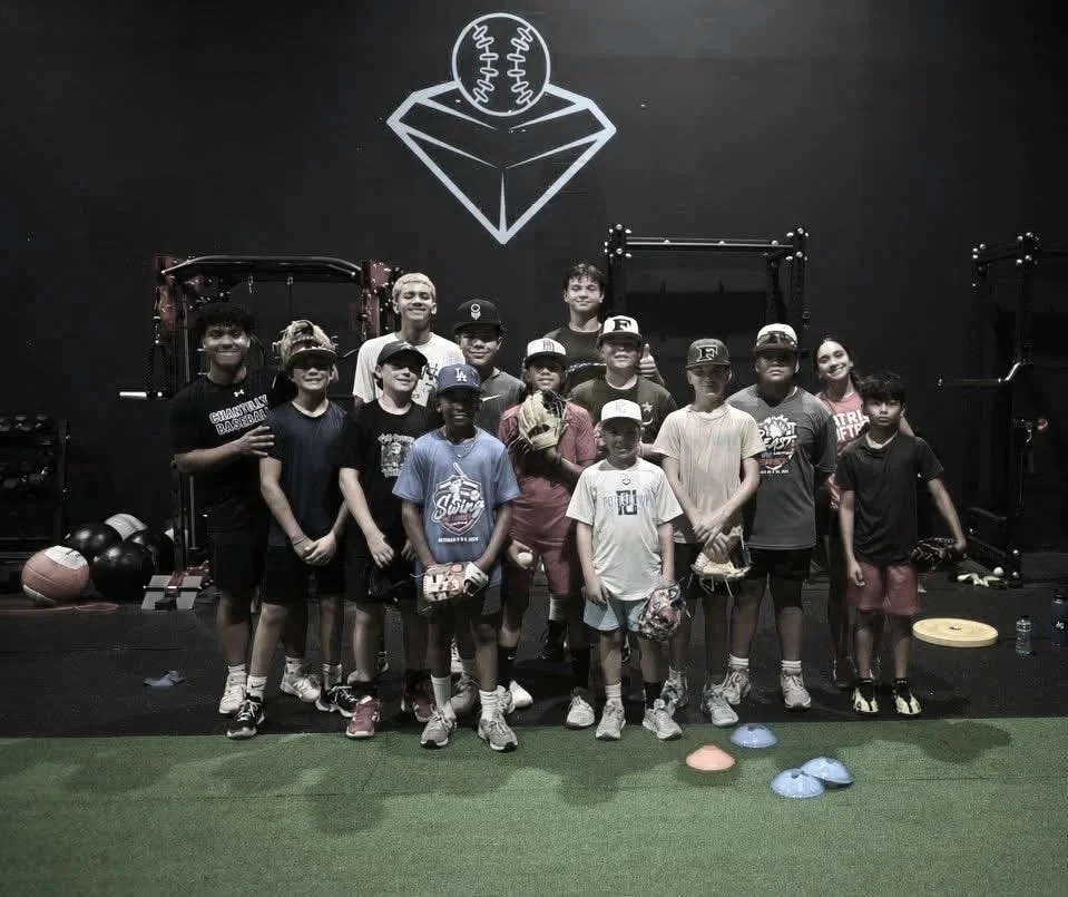 Group of children and teens in a gym with black walls, some with caps and sports gear, standing in front of gym equipment and baseball-themed artwork.