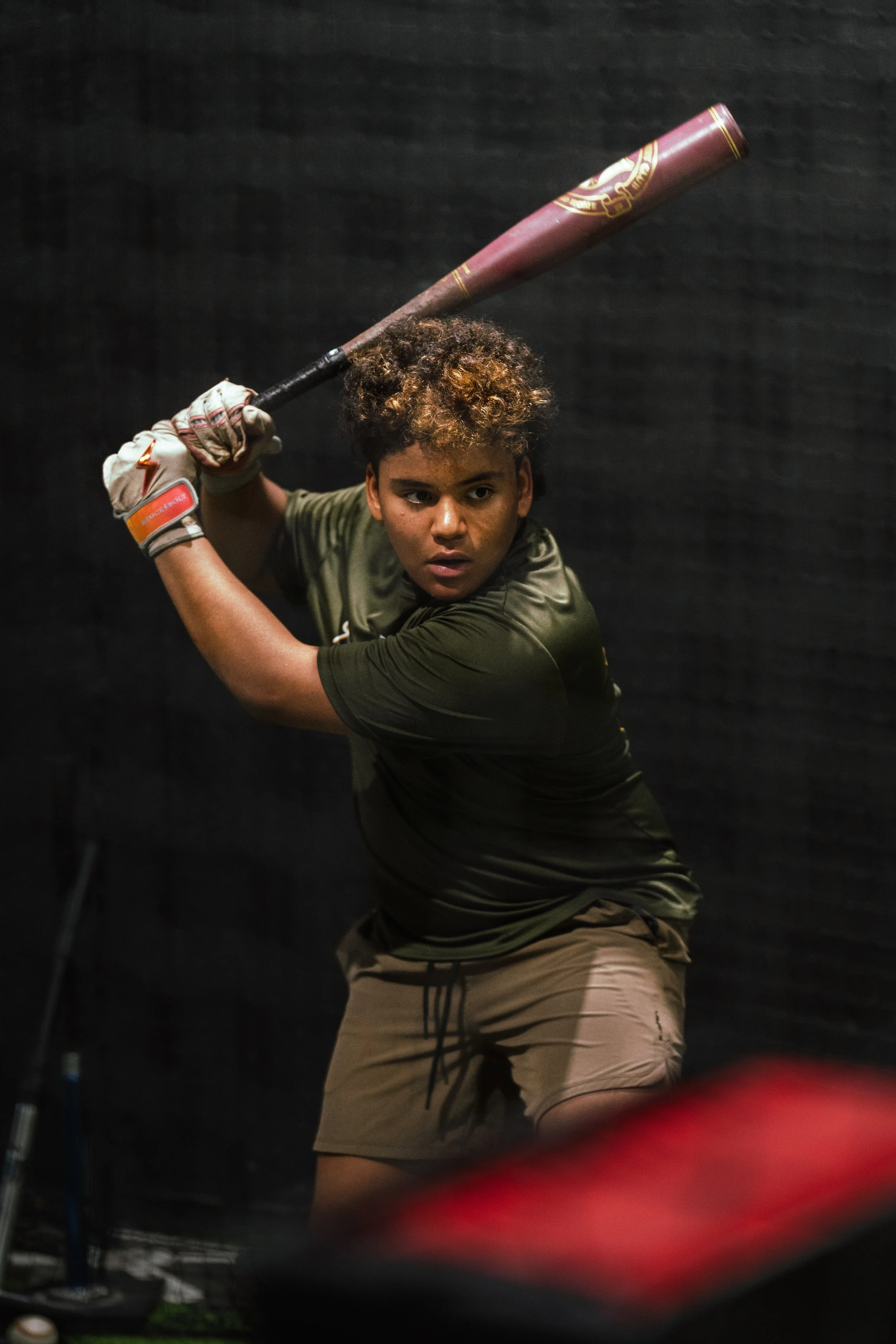 A young person with curly hair, wearing a green shirt and khaki shorts, is holding a baseball bat in a ready stance against a dark background.