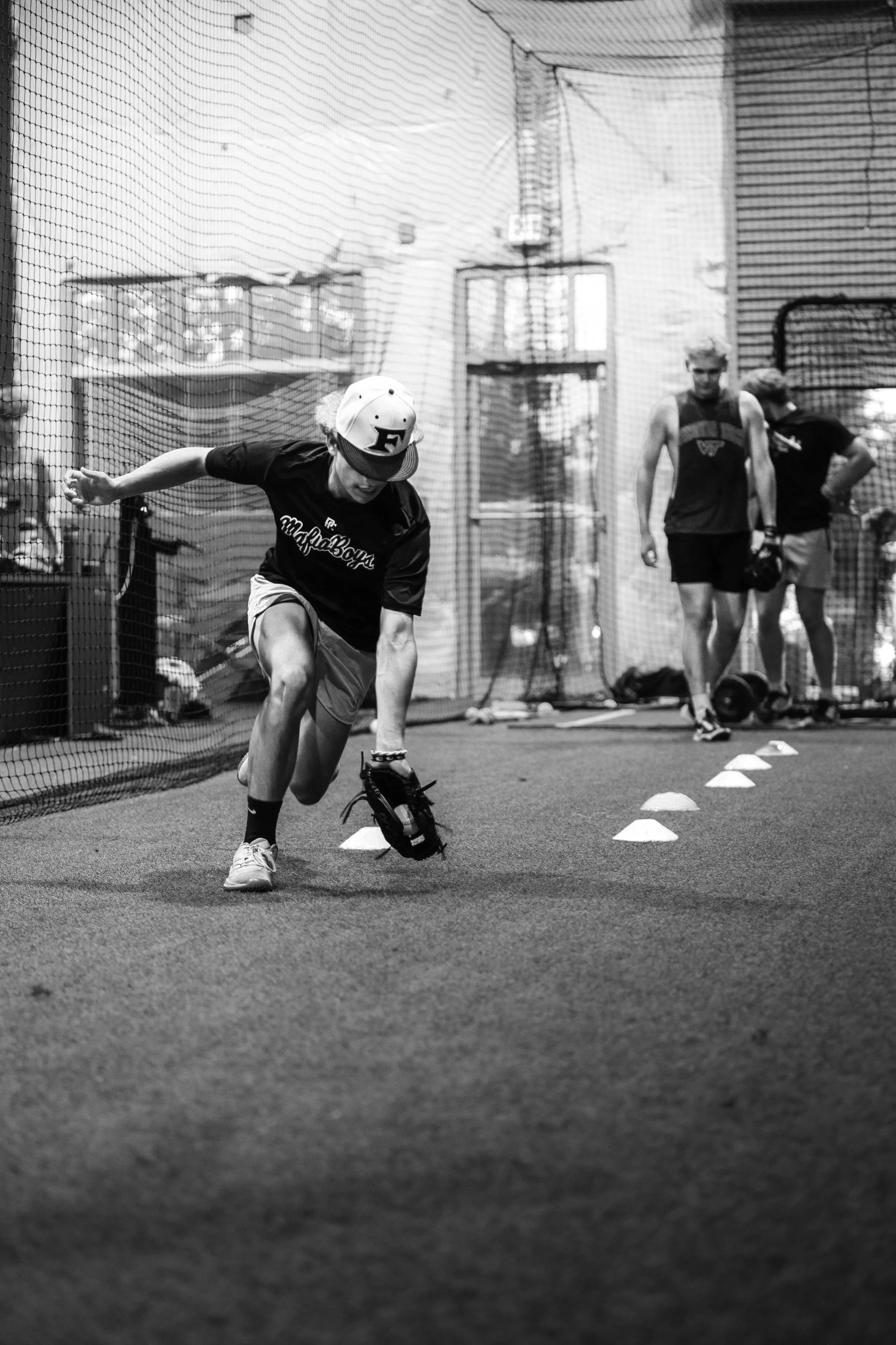 A young man wearing a baseball cap and athletic attire practicing baseball drills inside a gym, with two other athletes observing in the background.