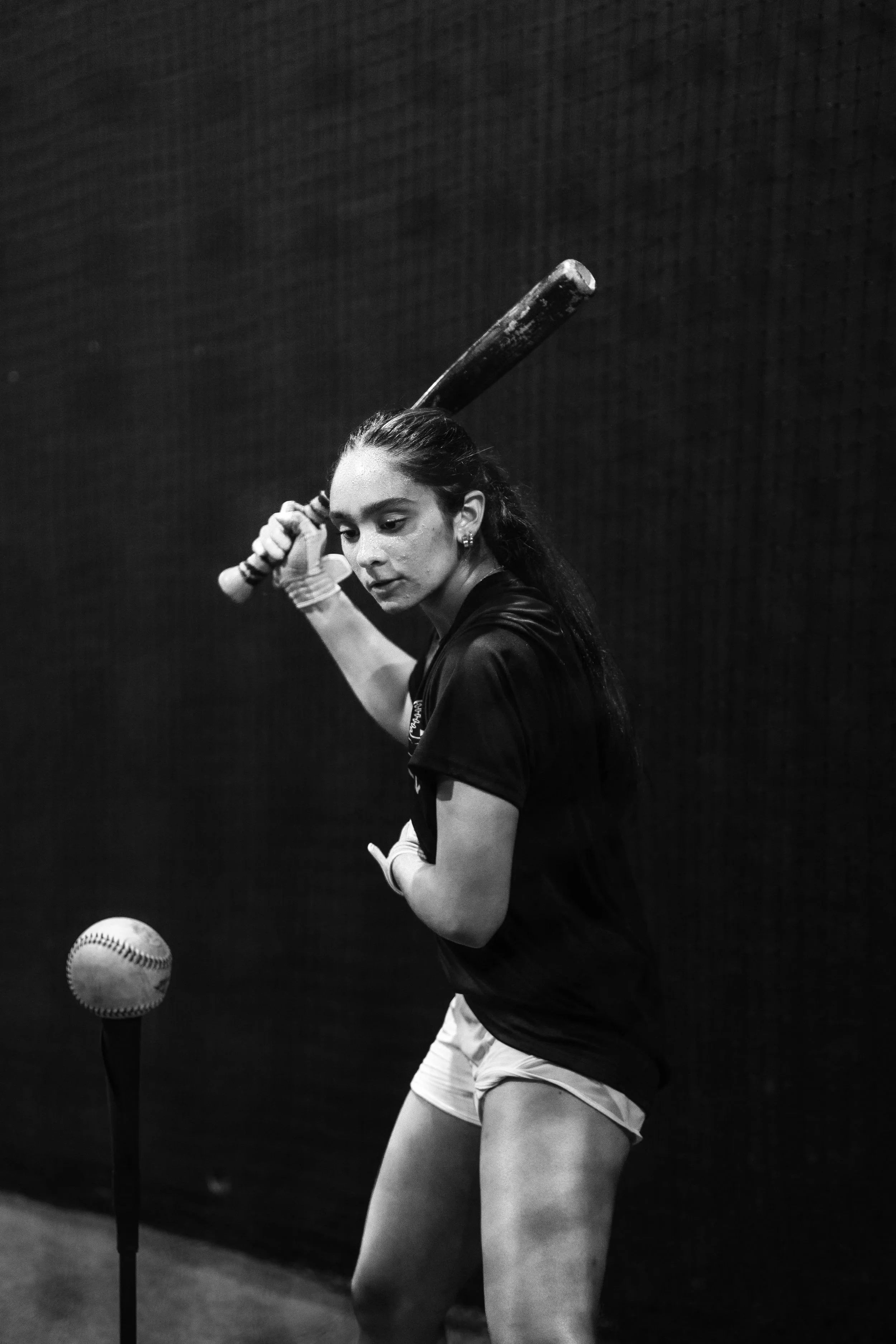 A young woman in sports attire holding a baseball bat, preparing to hit a baseball on a practice tee, with a focused expression.