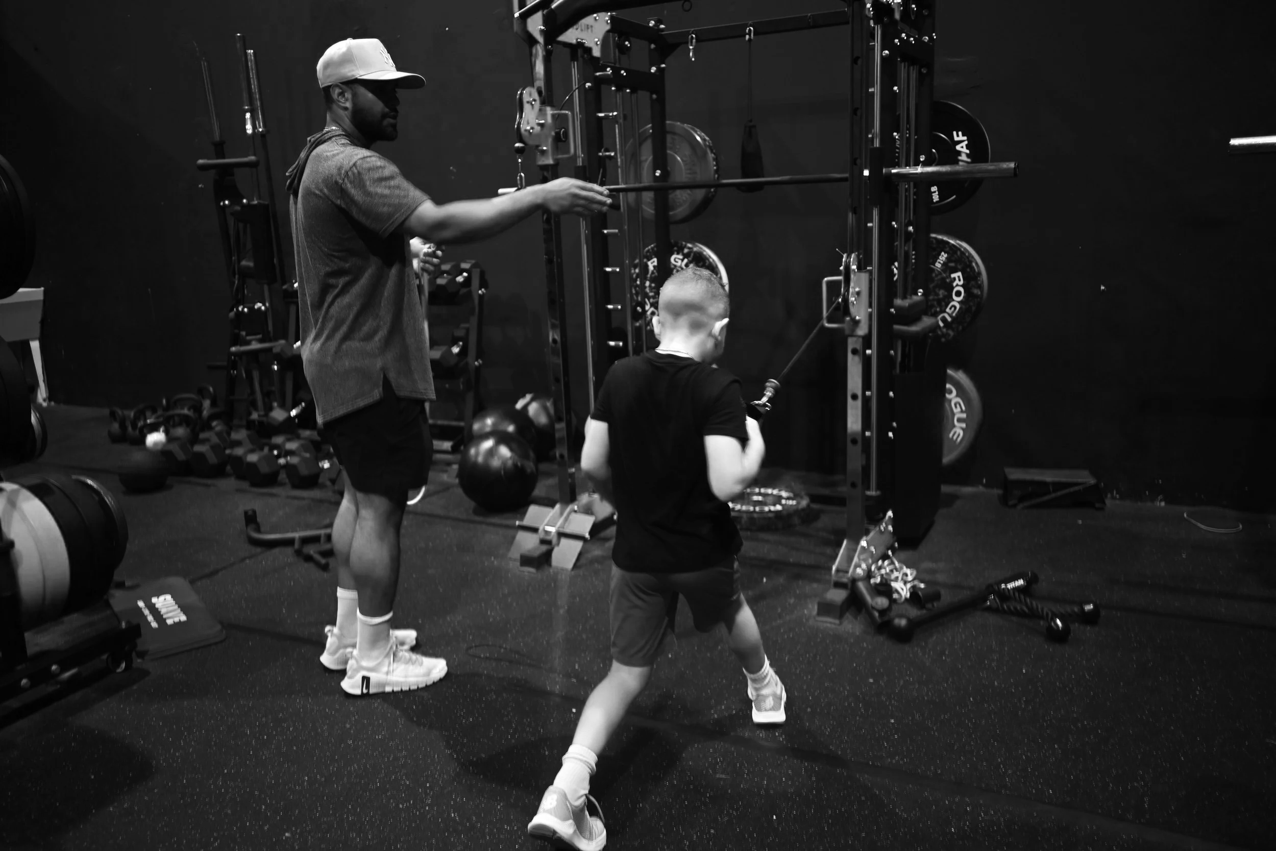 Black and white photo of a man and a boy working out in a gym. The man is standing and holding a barbell, while the boy is attached to a resistance machine, both focused on their exercise.