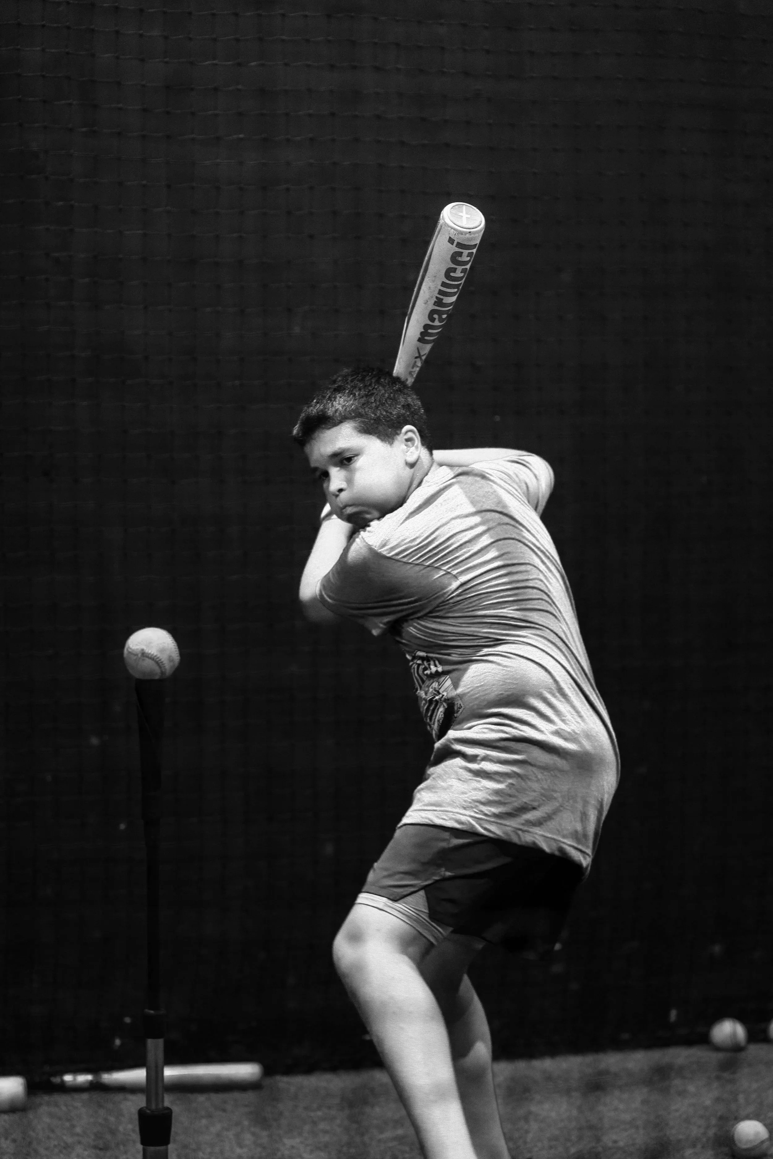 A boy playing baseball, holding a bat, preparing to hit a baseball pitched on a tee. The scene appears to be indoors with a dark background.