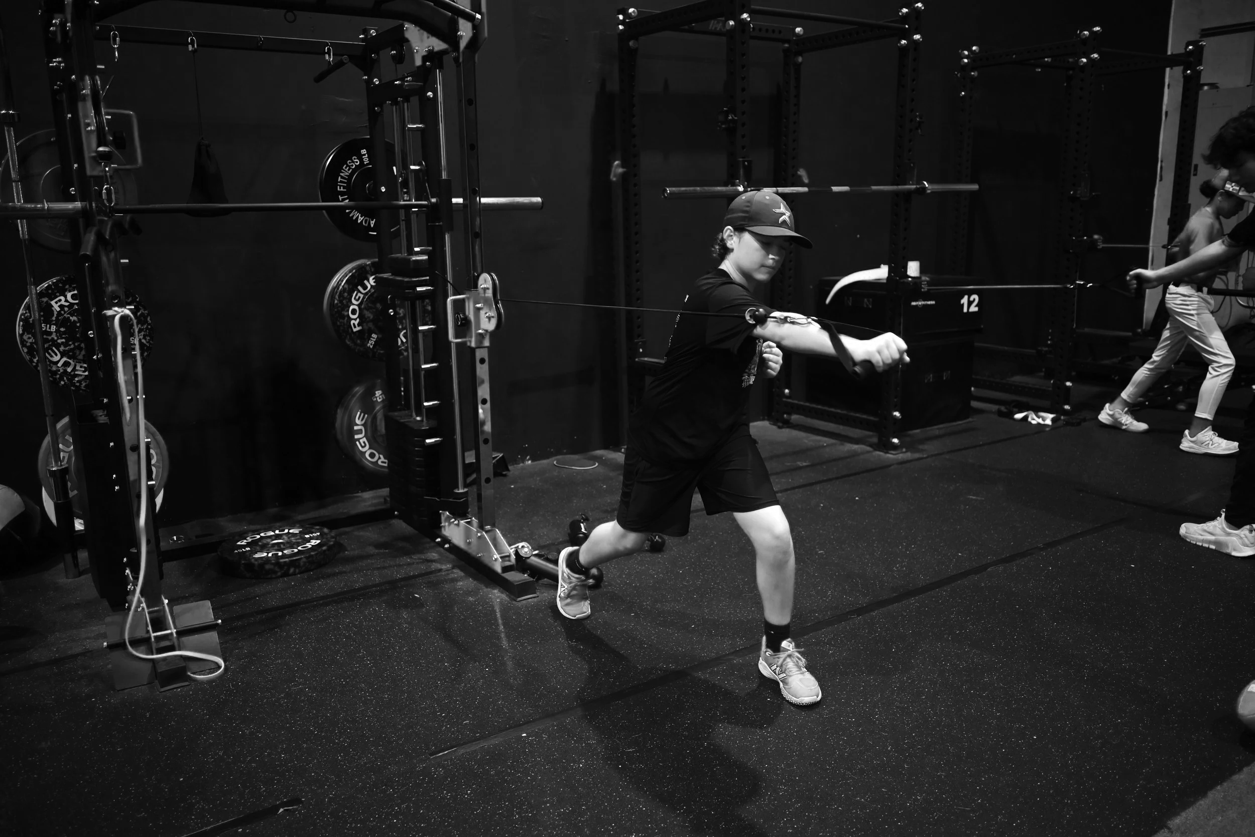 A young boy in a gym, wearing a baseball cap, T-shirt, shorts, and running shoes, is running while holding resistance bands, with gym equipment and other children in the background.