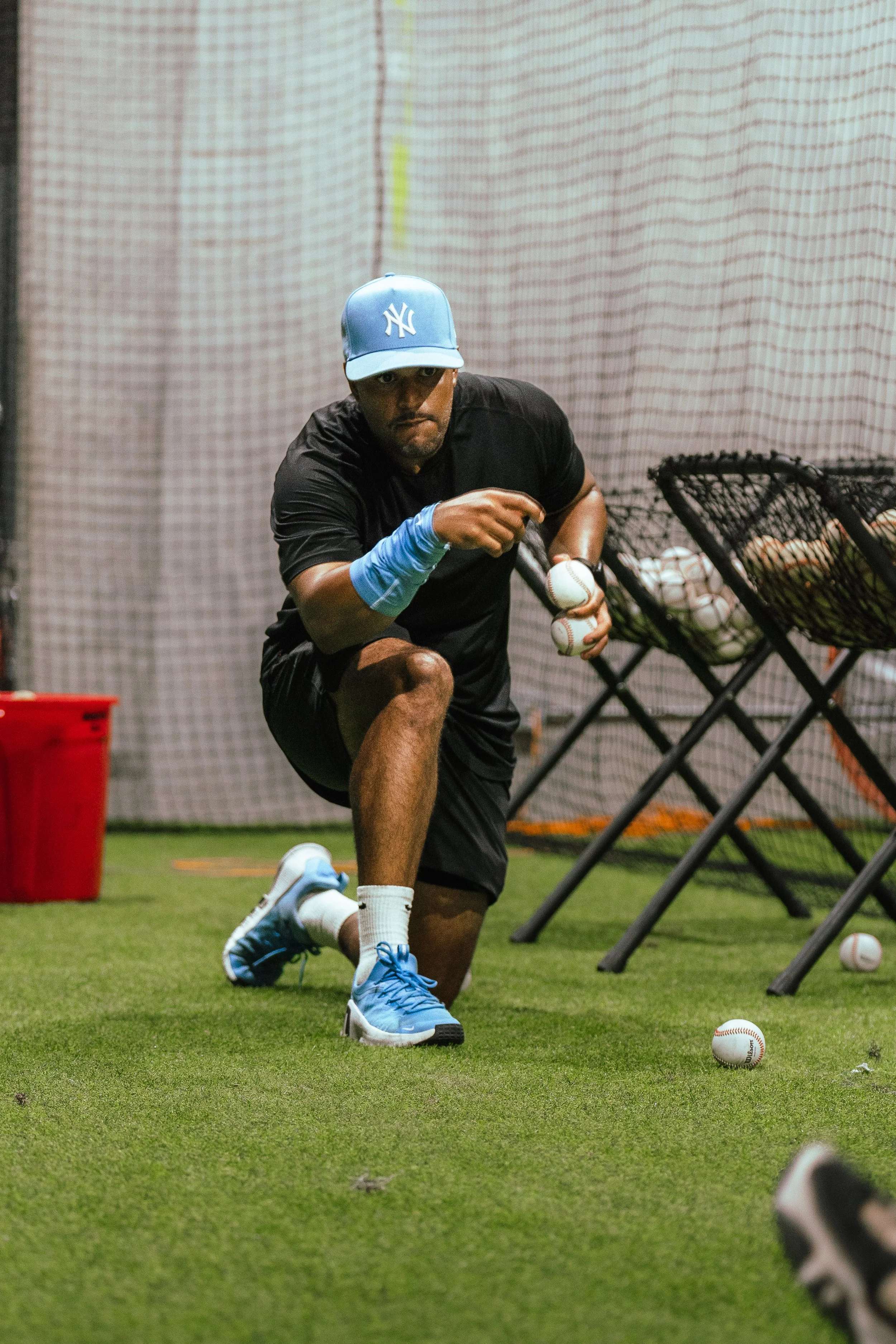 A baseball player is practicing his pitching indoors, wearing a black shirt, blue sneakers, and a light blue New York Yankees cap. He is holding baseballs in his left hand and is positioned in a crouched stance near a netted backdrop. There are multiple baseballs on the ground and in black netted baskets nearby.
