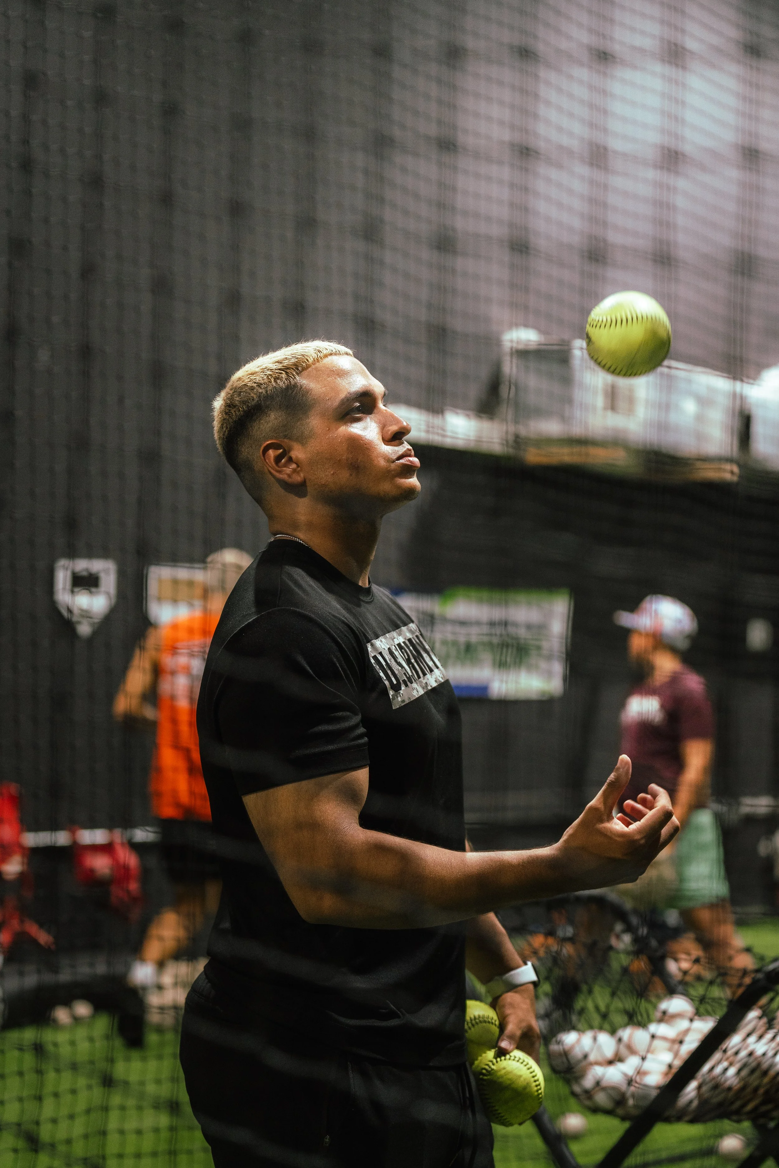 A young man with blond hair in a gym, wearing a black shirt, holding a softball in his right hand, under a net, with other people and equipment in the background.