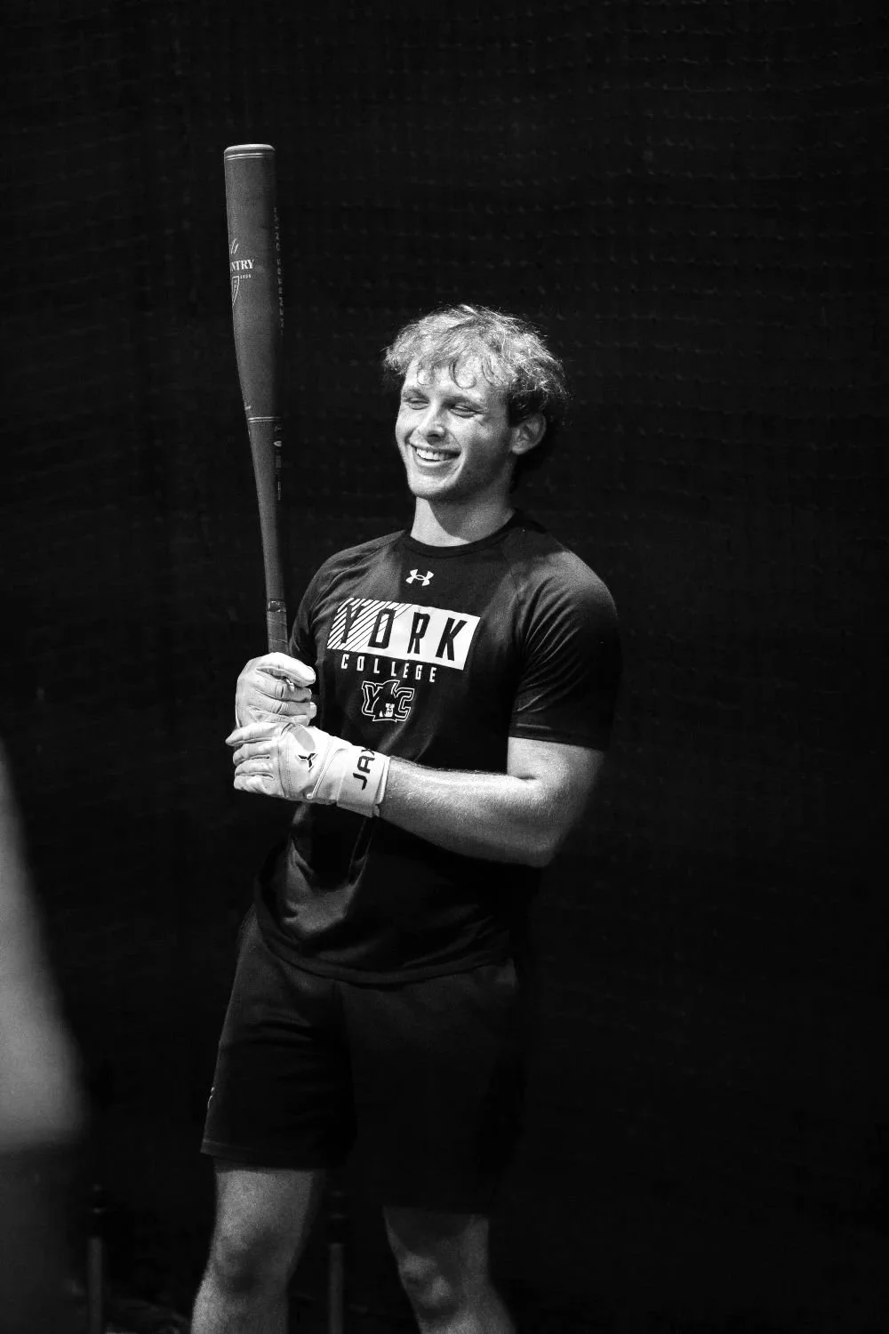 A young man with curly hair smiling, wearing a 'York College' athletic shirt and holding a baseball bat, standing against a dark background.