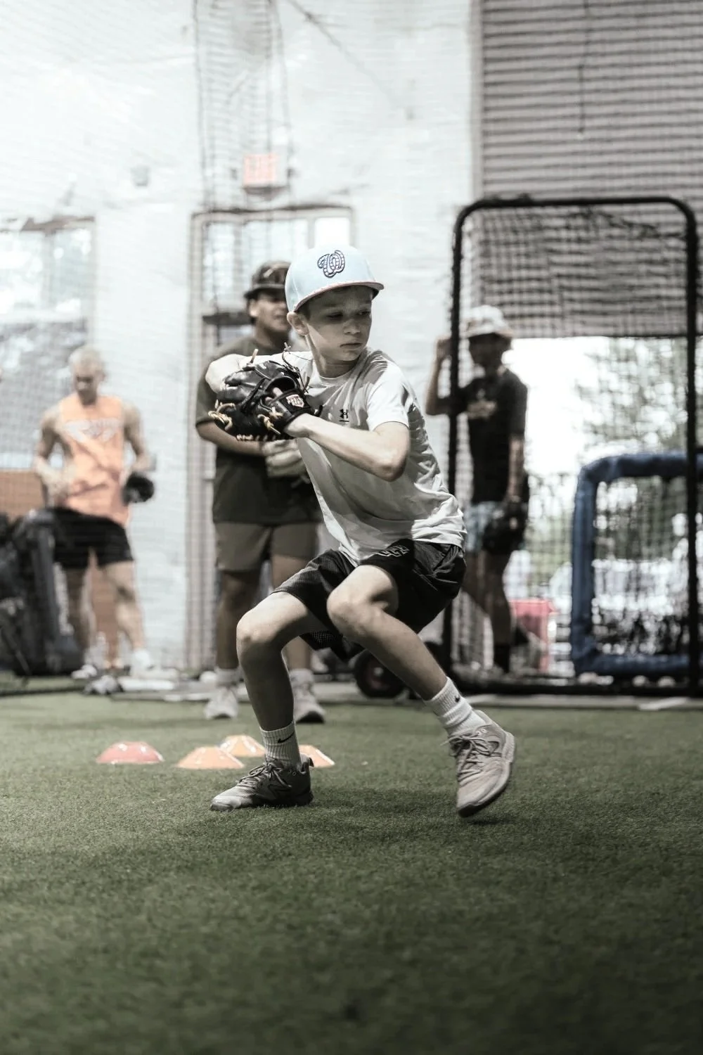 A young boy practicing baseball indoors, wearing a gray cap, a white shirt, and black shorts, holding a baseball glove, with coaches and other children watching in the background.