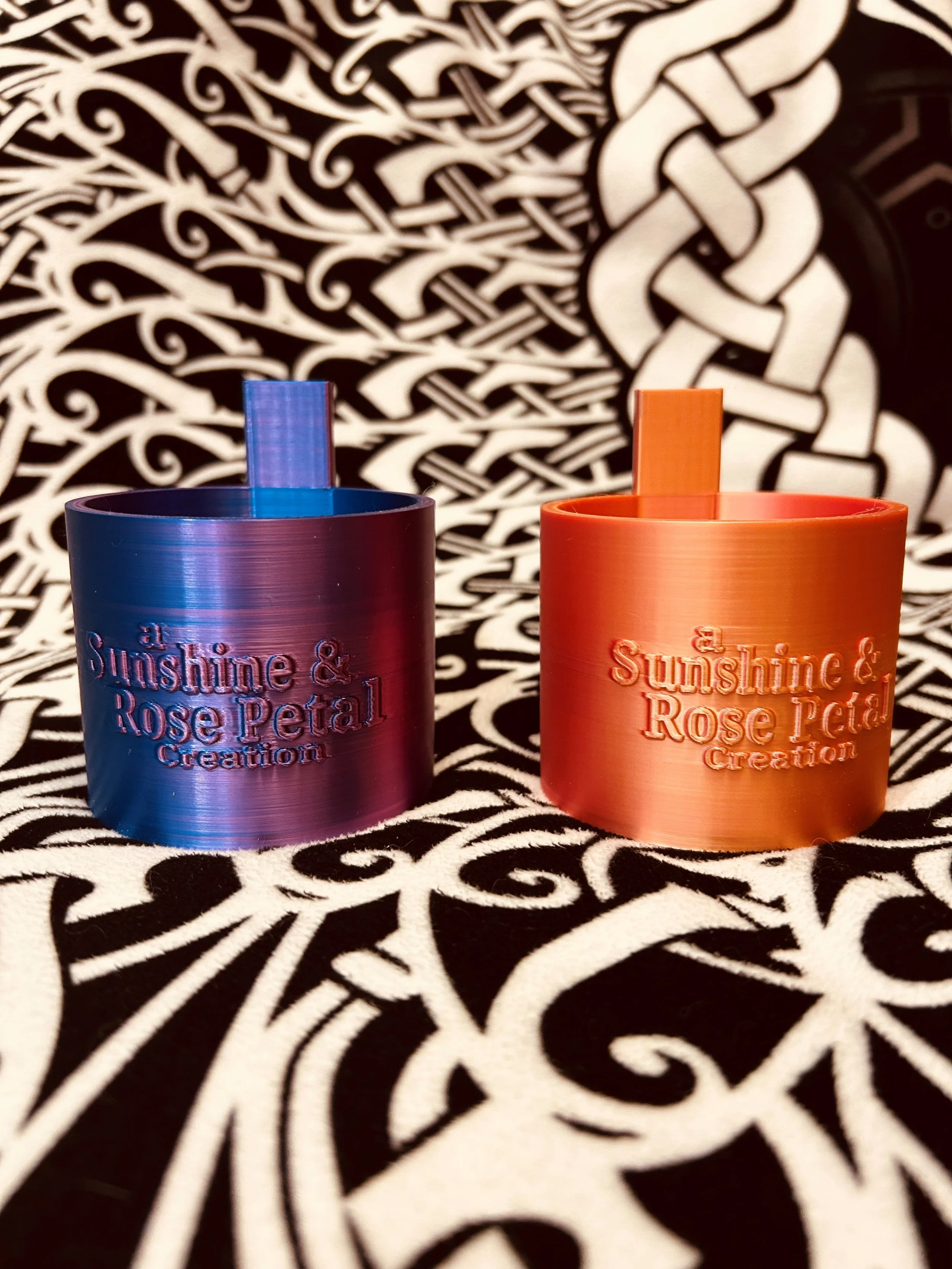 Two metallic bottles with engraved text, one blue and one orange, placed on a black and white patterned fabric. The blue bottle is on the left, and the orange bottle is on the right.