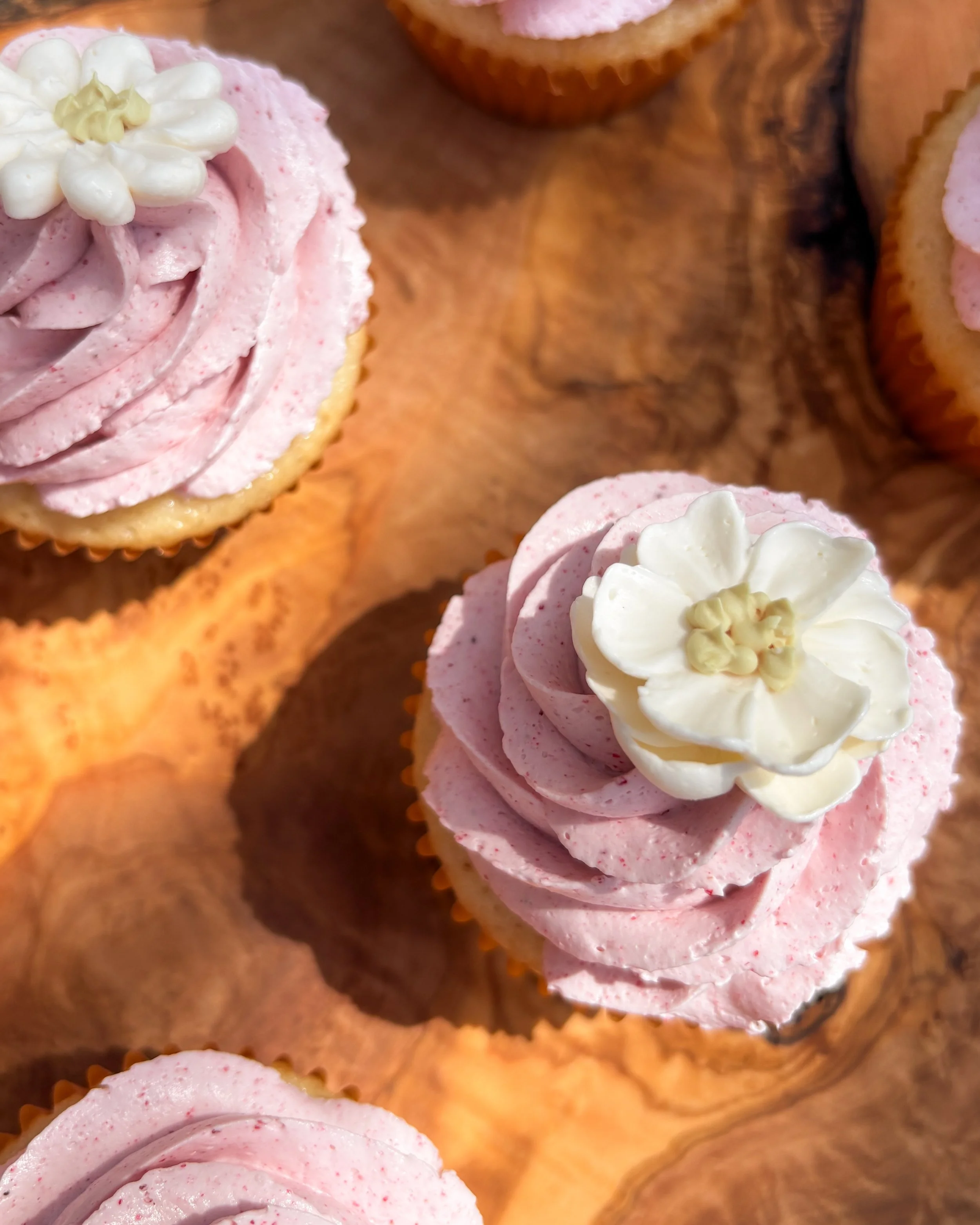Close-up of pink cupcakes with white flower-shaped icing decorations on top, placed on a wooden surface.