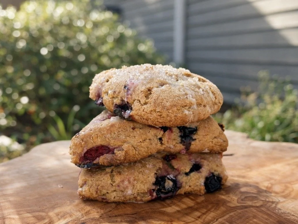 Three freshly baked berry lemon scones stacked on a wooden surface outdoors, with a blurred background of greenery and a house.