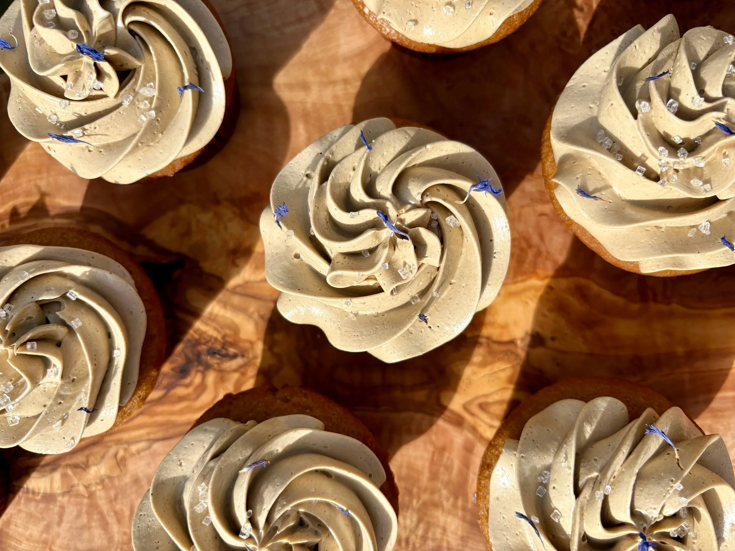 Close-up of cupcakes with light brown frosting piped in rose shapes, decorated with edible blue flowers and small white salt crystals, on a wooden surface.