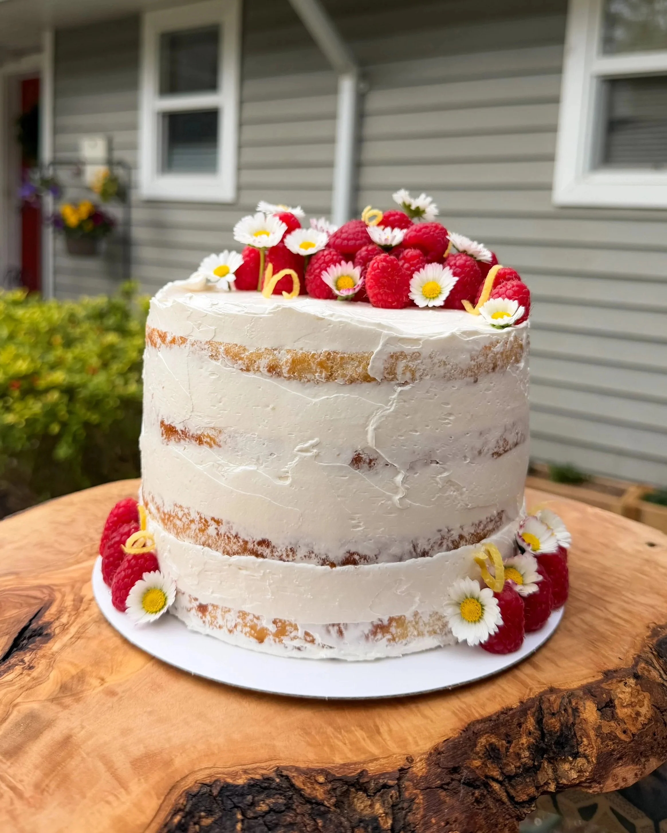 A three-tiered cake decorated with fresh raspberries, daisies, and lemon zest on a wooden cake stand outdoors near a house with grey siding and white-framed windows.