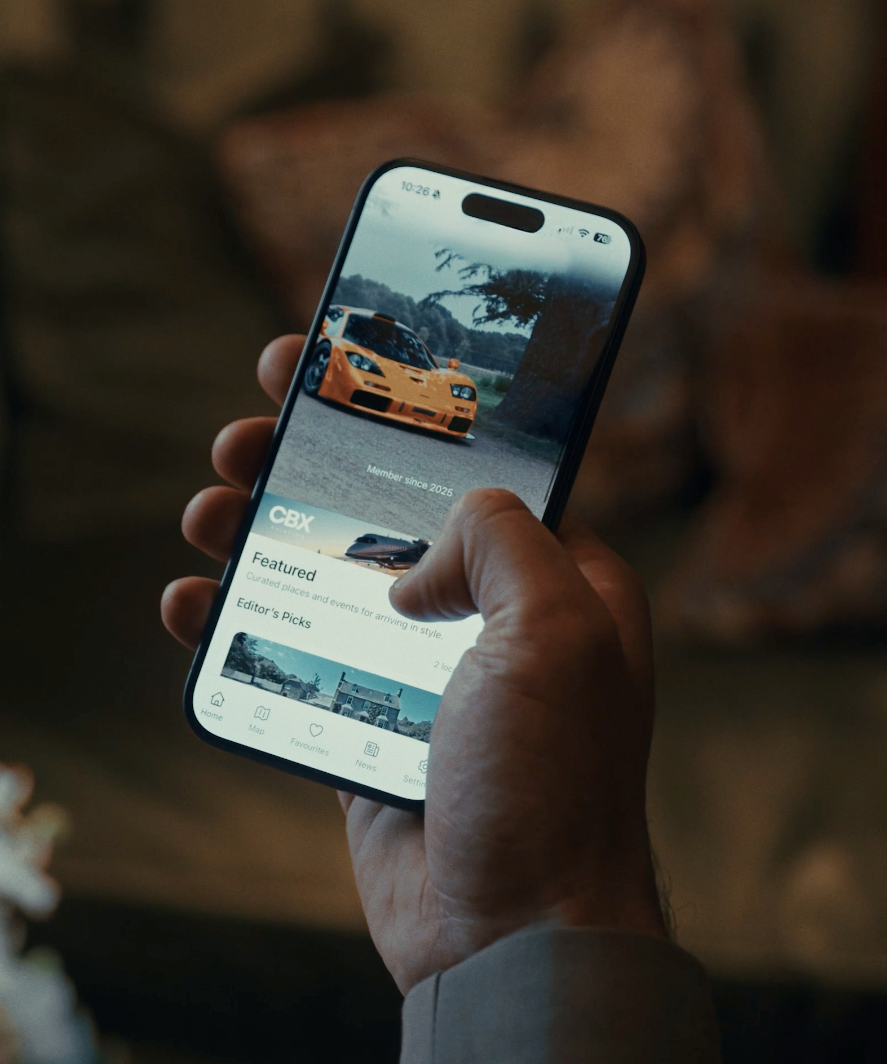 Close-up of a hand holding a smartphone displaying a car-themed app. The screen shows a yellow sports car on a scenic background, with menu options at the bottom and text indicating membership since 2025.