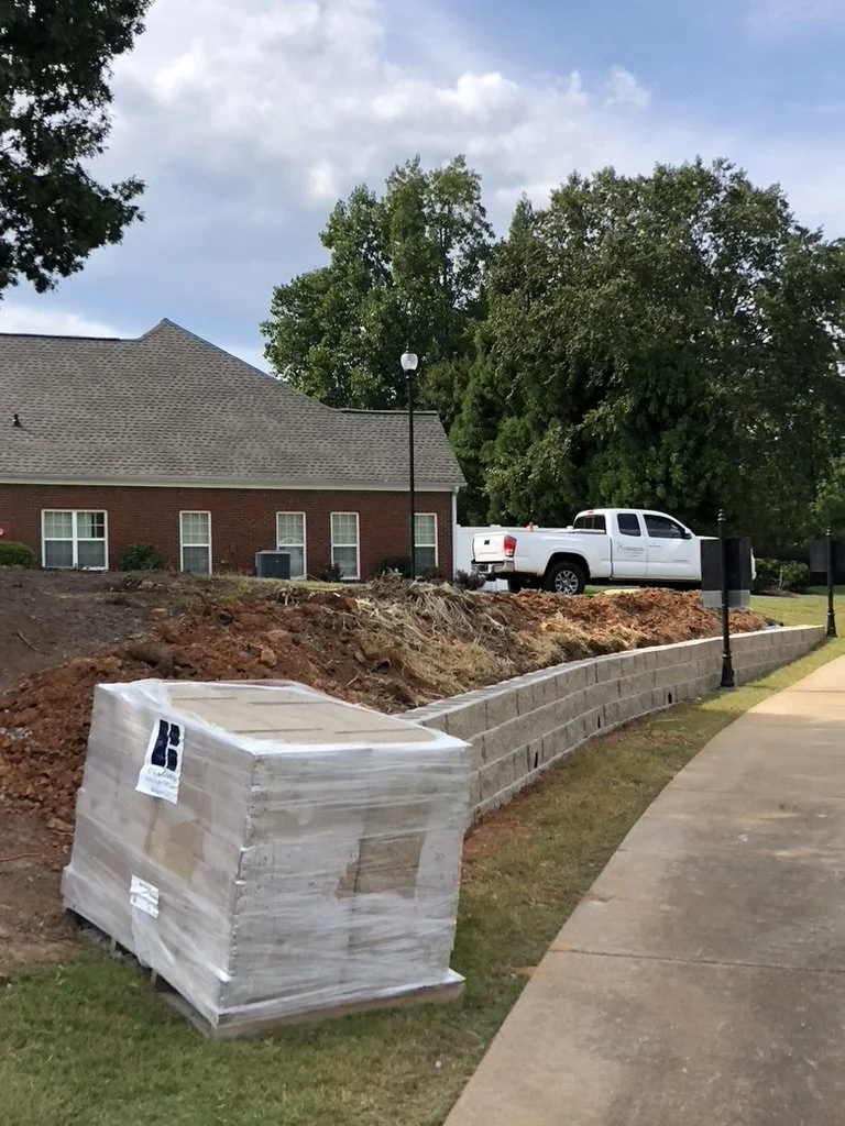 Sidewalk and ground dig-up with a retaining wall, construction materials, and a pickup truck with trees in the background.