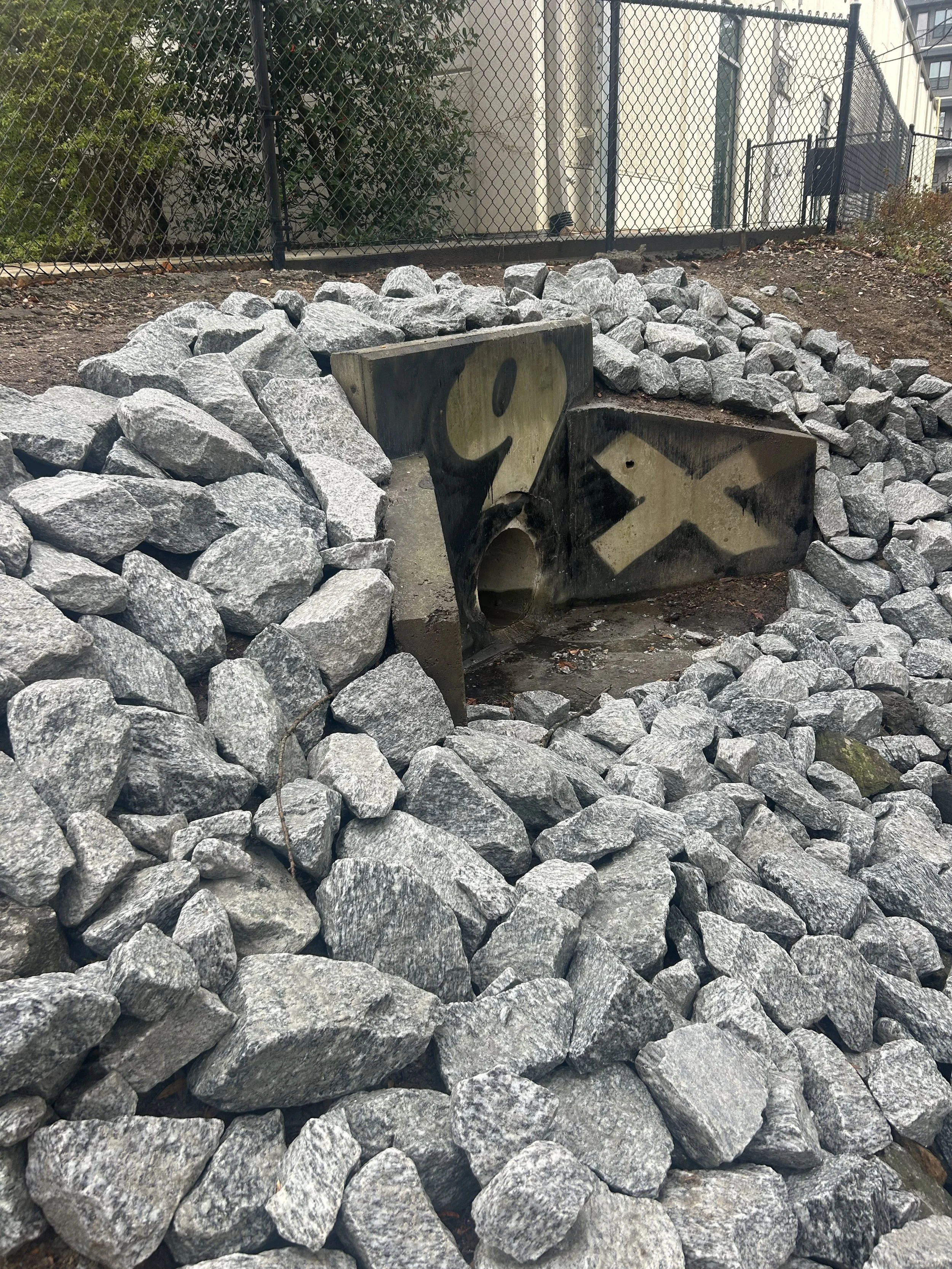A rock garden with a concrete piece featuring graffiti, surrounded by large gray rocks, with a black chain-link fence and buildings in the background.