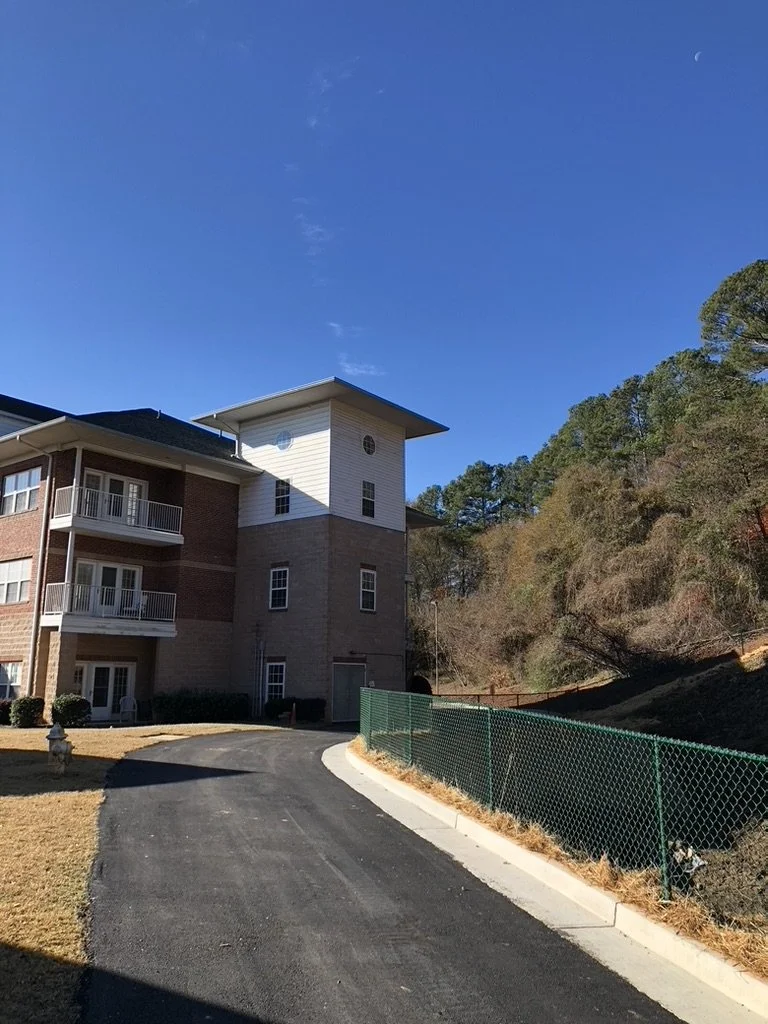 A residential apartment building with brick and white siding, multiple balconies, and a small tower with circular windows, situated next to a paved road, green fence, and wooded hillside under a clear blue sky.