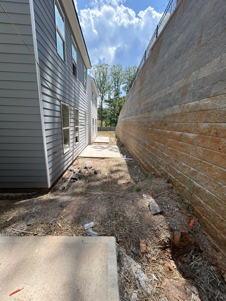 A narrow backyard between a gray house with horizontal siding and a tall wooden privacy fence under a partly cloudy sky.