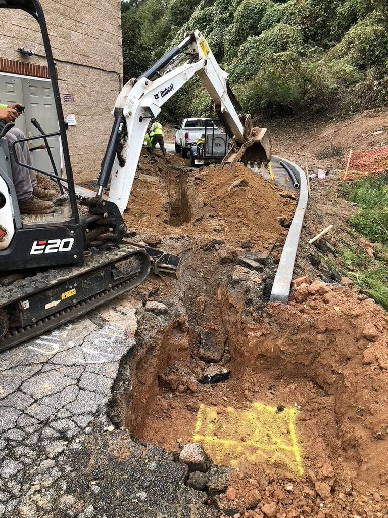 Construction workers repairing a damaged road with an excavator digging a trench along the sidewalk next to a building, with a white vehicle and a trailer in the background.