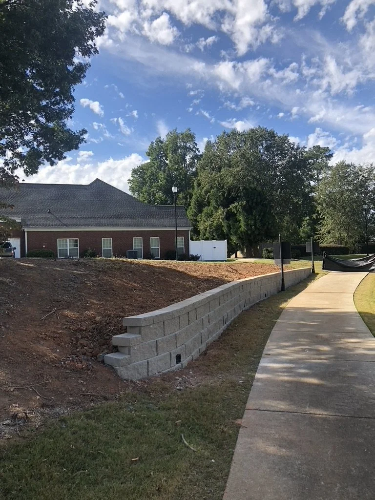 A curved stone retaining wall with a beige sidewalk next to it, grass on the other side, and a brick building with several windows in the background, under a partly cloudy sky.