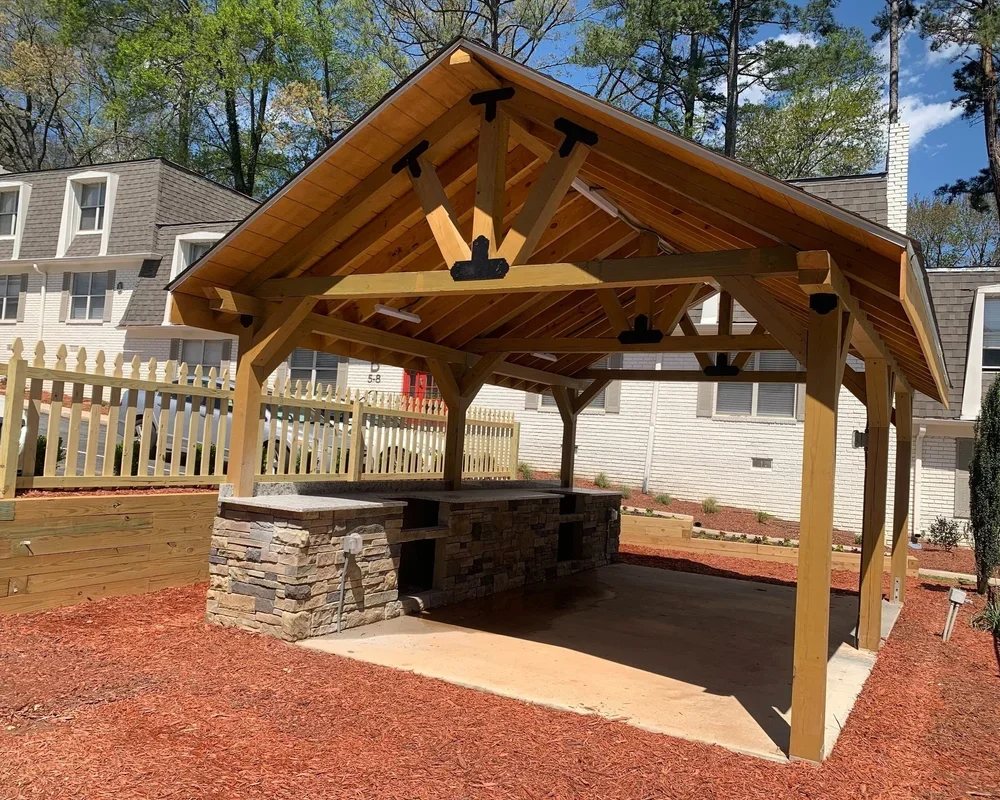Wooden pavilion with a stone base, situated in a yard with red mulch and a wooden fence in the background, in front of white houses and trees.