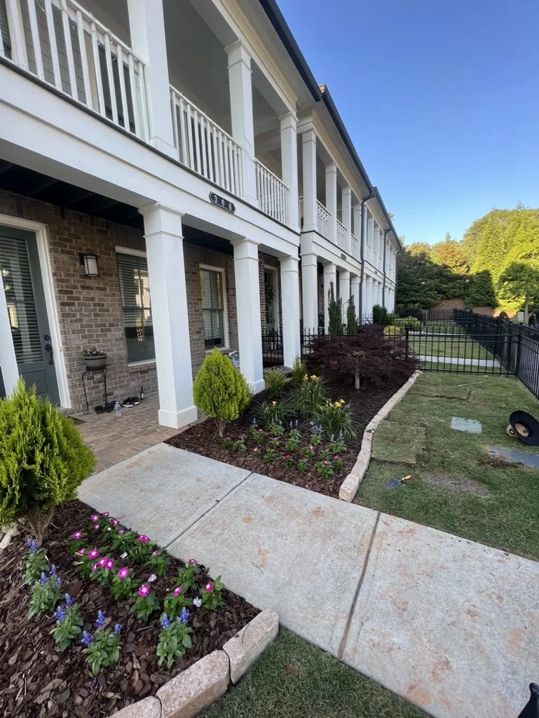 Exterior view of a two-story residential building with white pillars, brick walls, and a balcony. There are landscaped garden beds with flowers, shrubs, and small trees, along with a concrete walkway and a grassy area with a black fence.