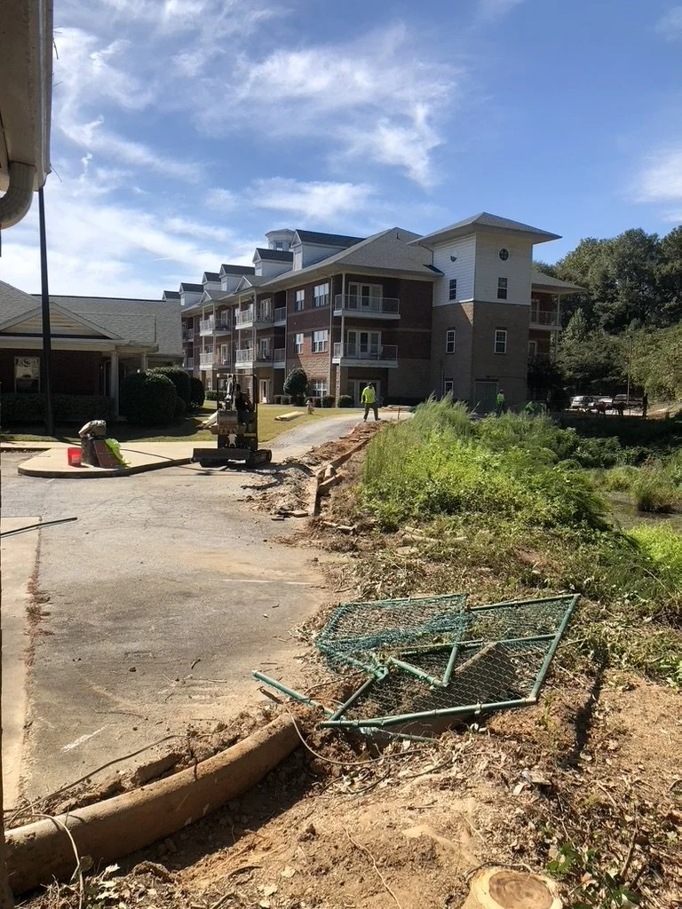 Construction workers working on a path near a multi-story residential building, with a fallen green metal fence in the foreground and a partly cloudy sky above.