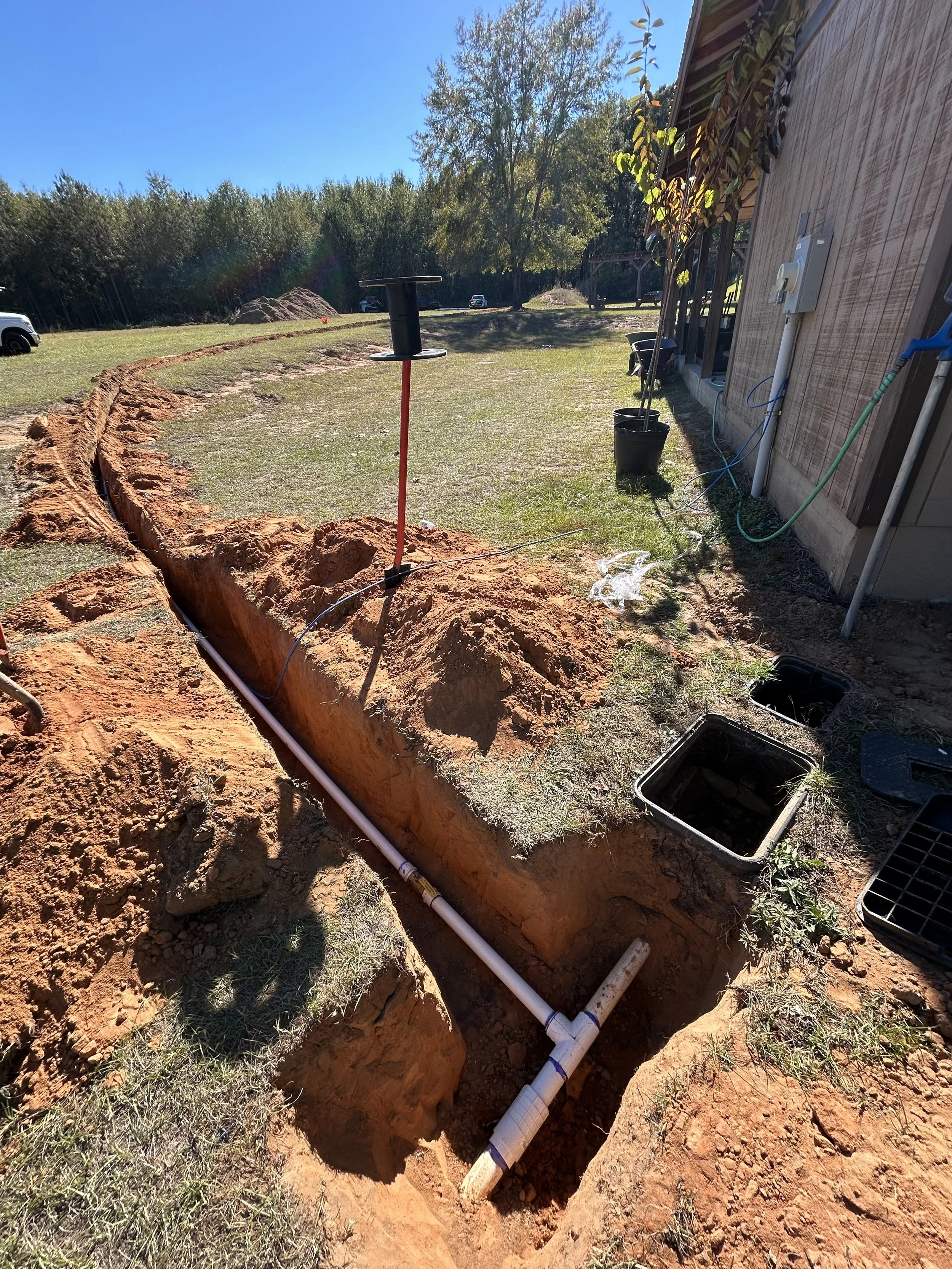 Exposed underground plumbing with PVC pipes near a building, with a trench dug for installation, on a sunny day with green grass and trees in the background.