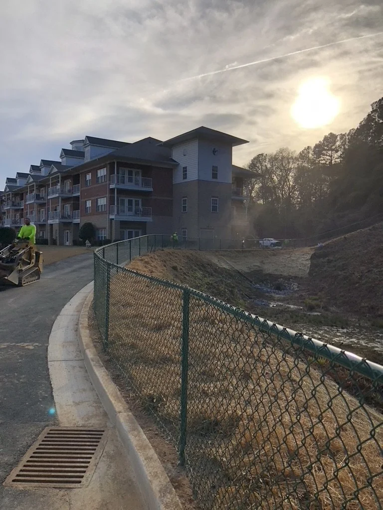 Construction site with a partially finished multi-story apartment building, a chain-link fence, and a worker operating a compact skid-steer loader, with the sun low in the sky behind trees.