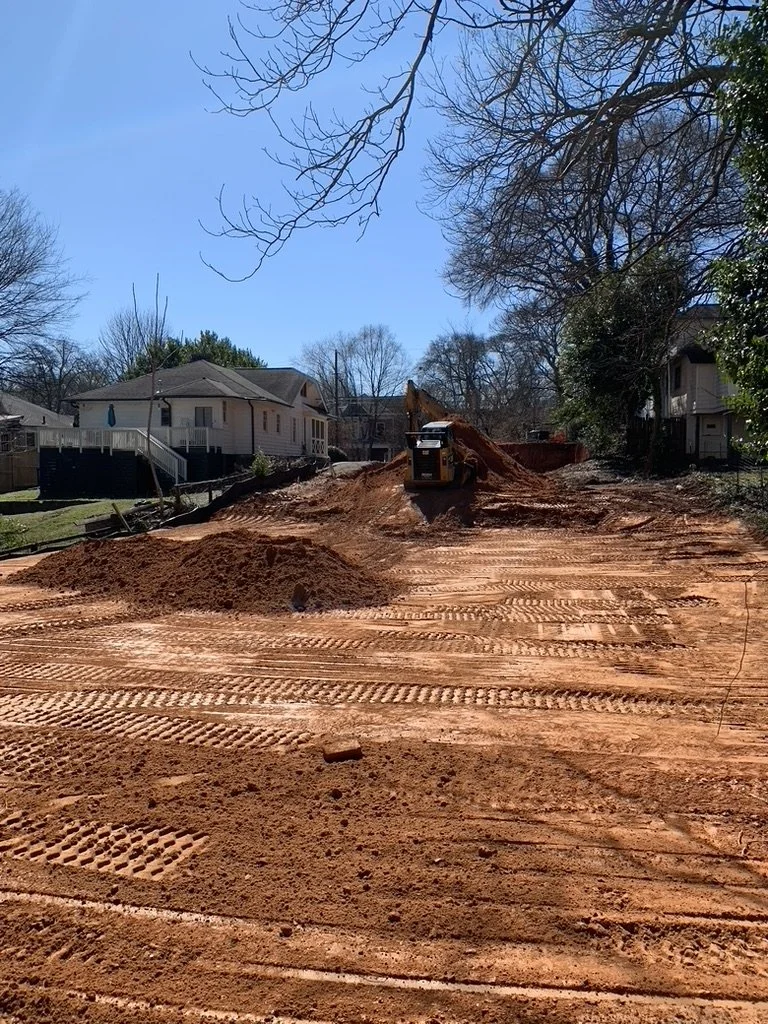 Construction site with exposed dirt, tire tracks, and an excavator, surrounded by residential houses and trees.
