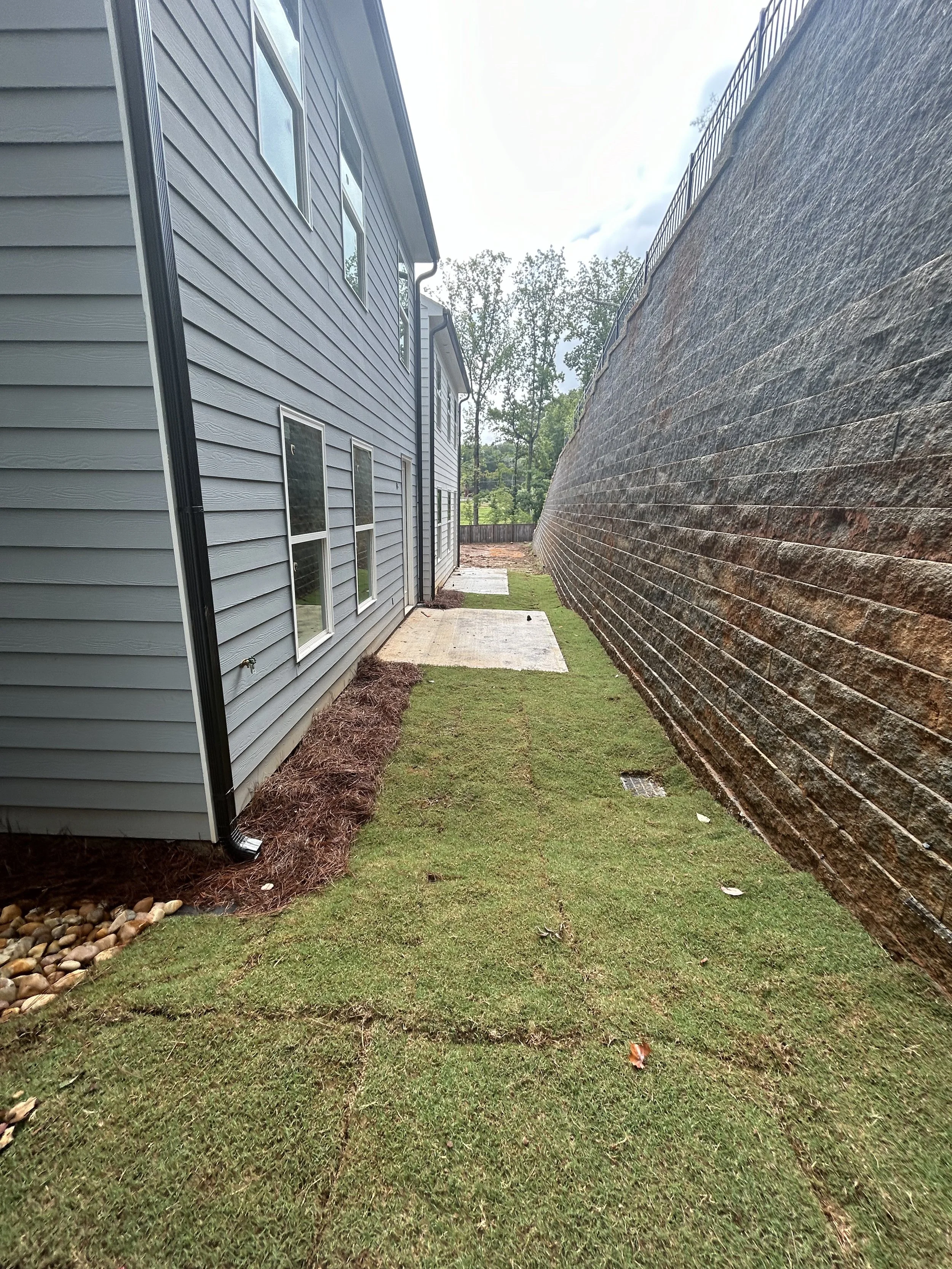 A side yard between a gray house with three windows and a tall brick wall, with patches of grass and concrete slabs on the ground.