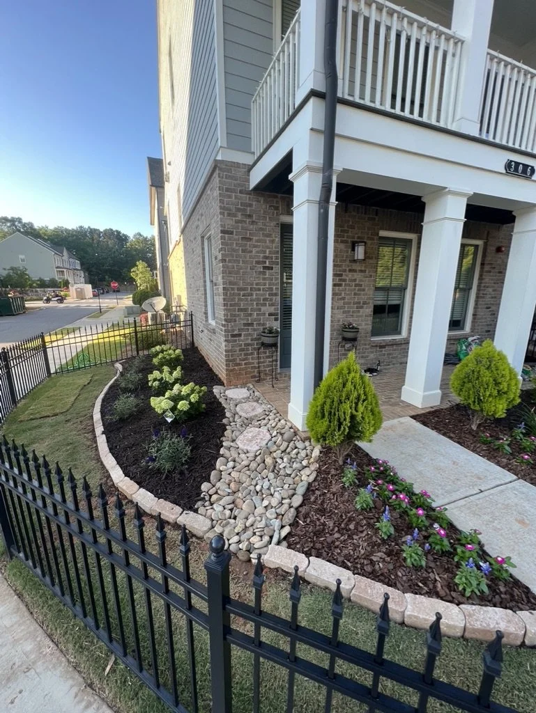Front porch garden with small shrubs, flowers, a stone pathway, and a black fence.