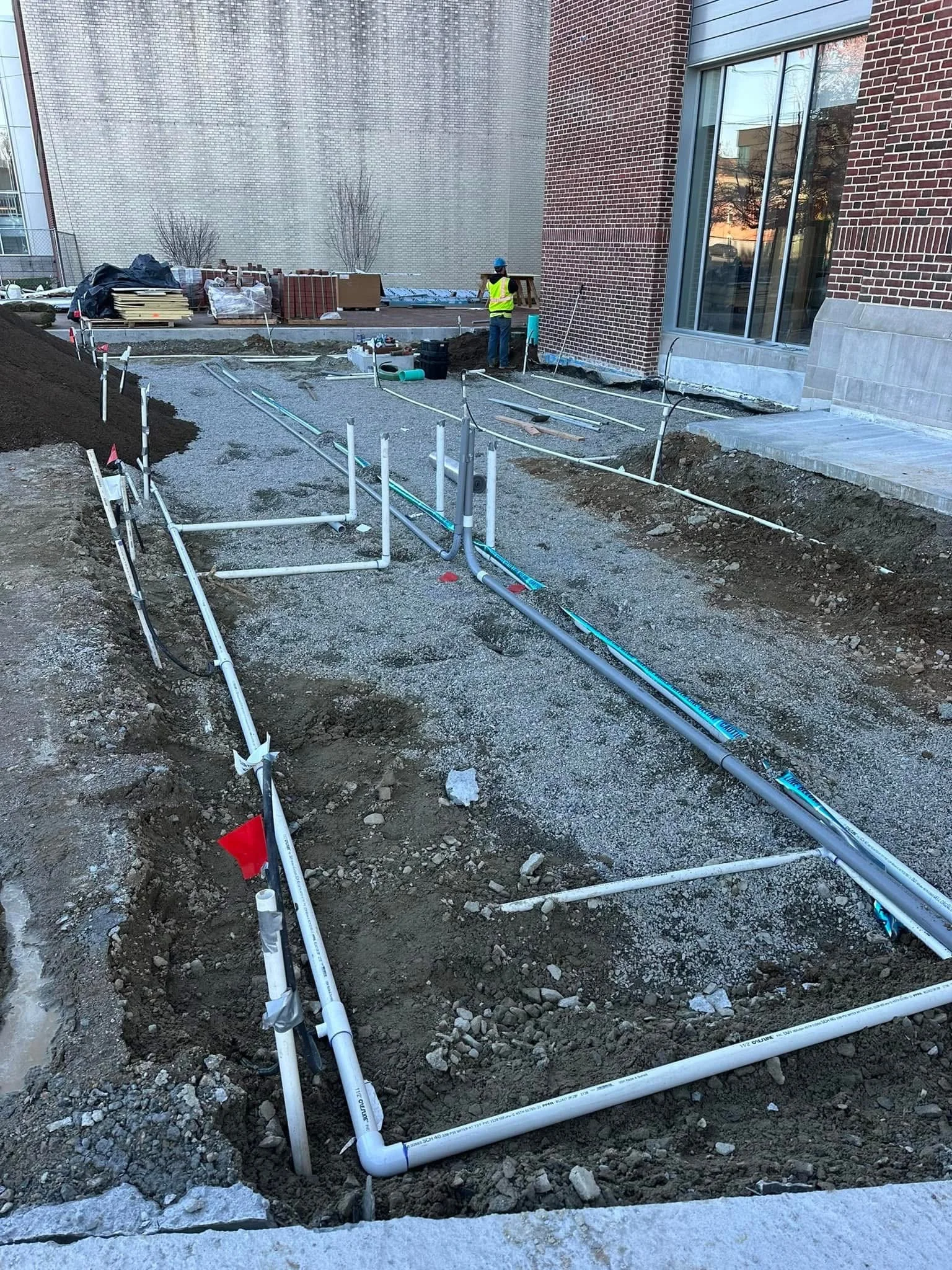 Construction site with exposed piping and two workers wearing safety vests and helmets working near a building wall.