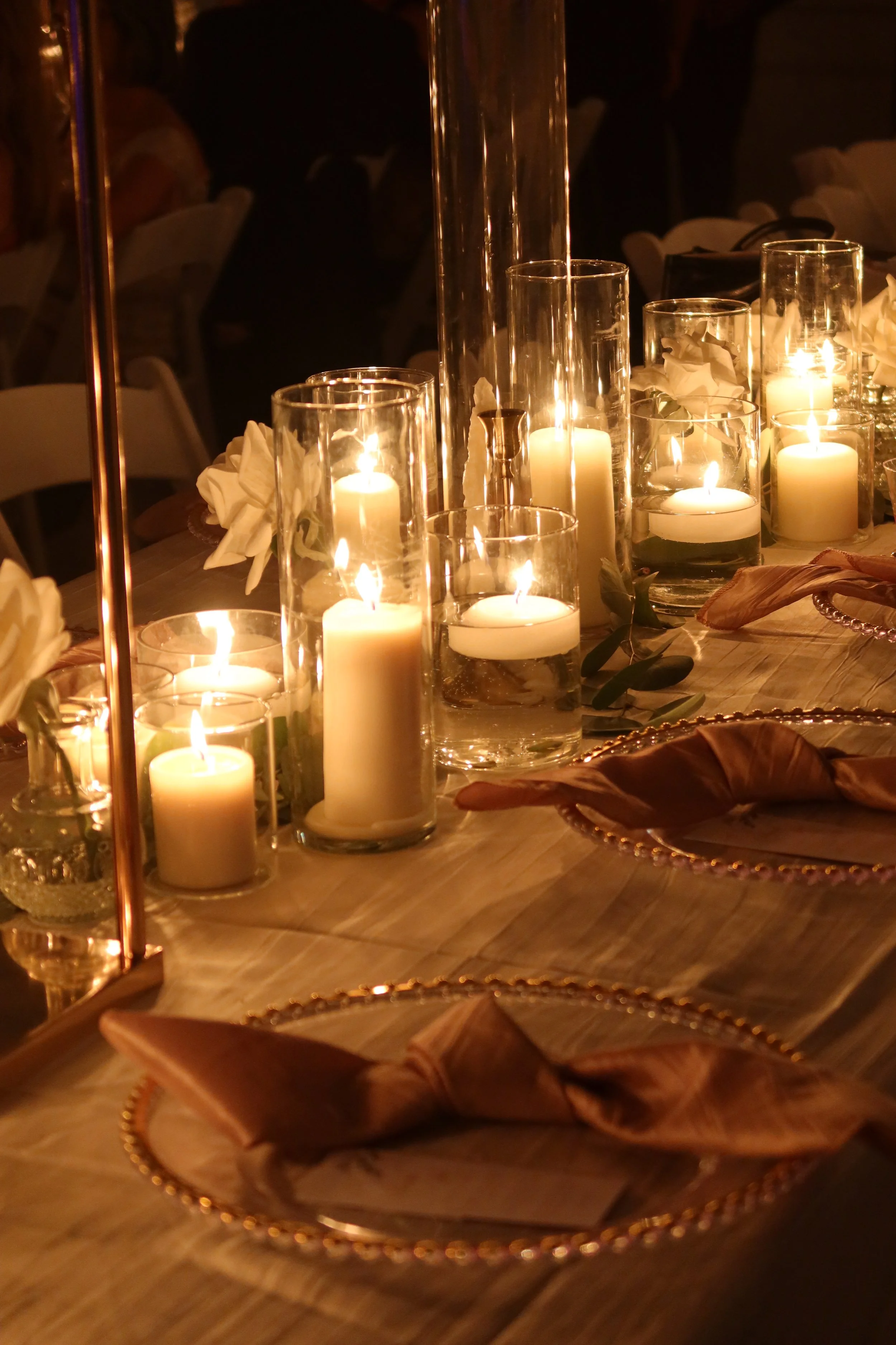 Elegant dining table decorated with candles in glass holders, surrounded by cloth napkins and floral centerpieces.