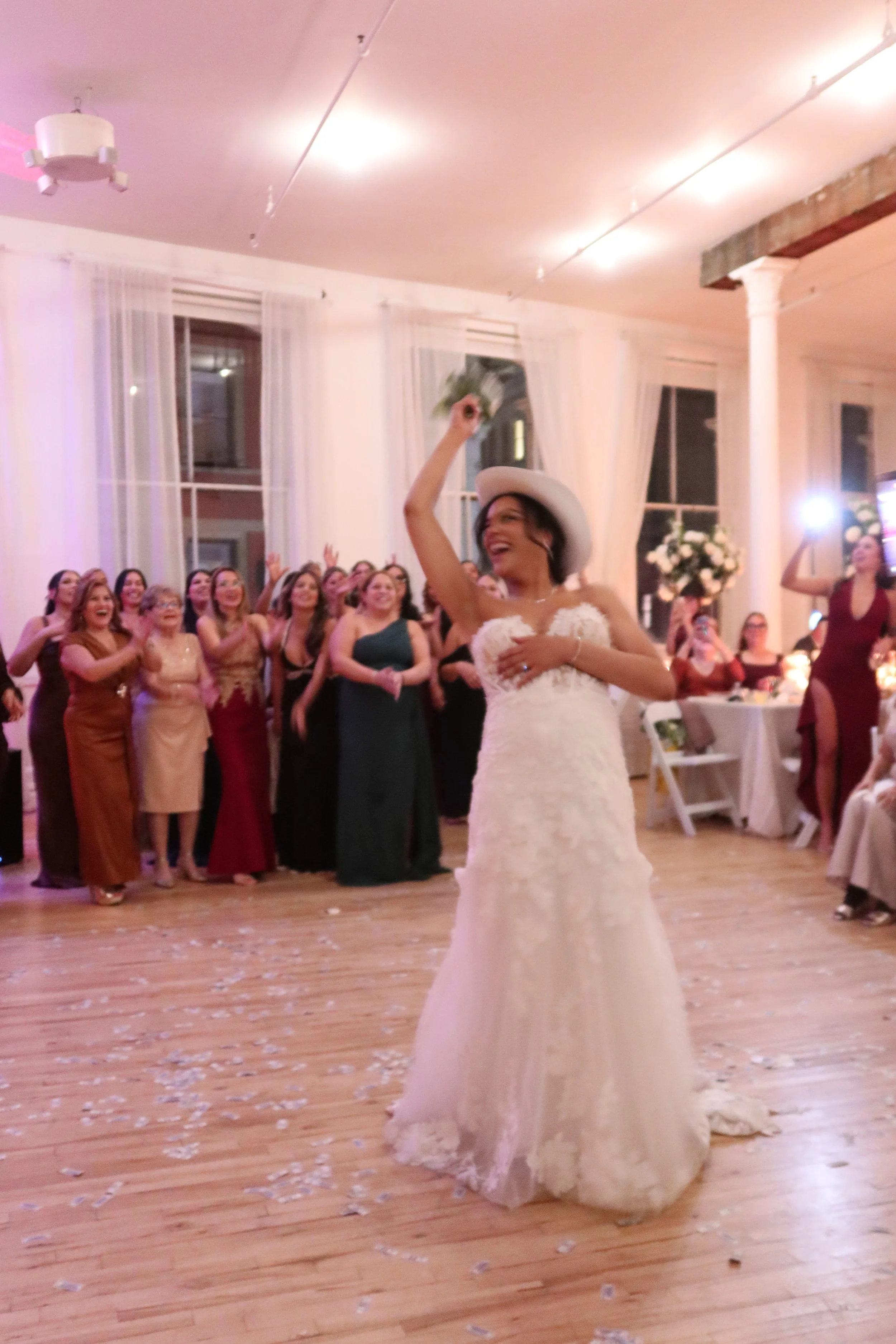 A bride in a white wedding dress holding a bouquet and wearing a white hat, celebrating with her wedding guests in an indoor reception hall. The bride is smiling and surrounded by women clapping and cheering.
